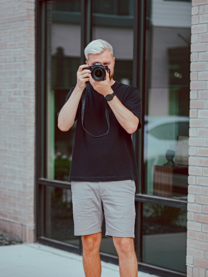 A social media manager with blonde hair taking a photo with a camera outside a modern building with large windows and brick walls.