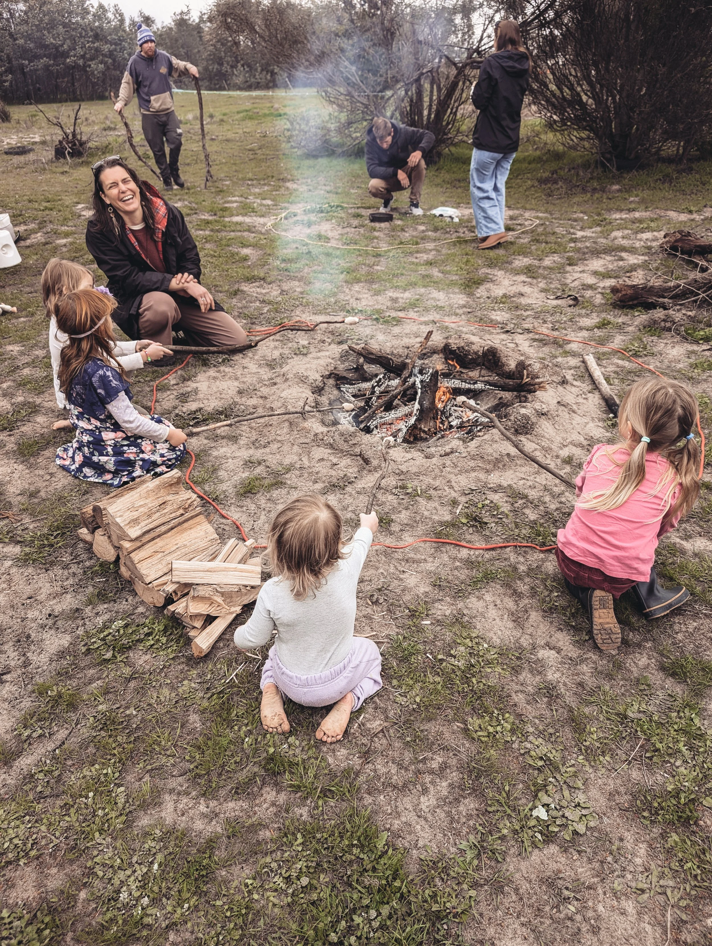 Children roasting marshmallows around a campfire with adults nearby on a grassy field.