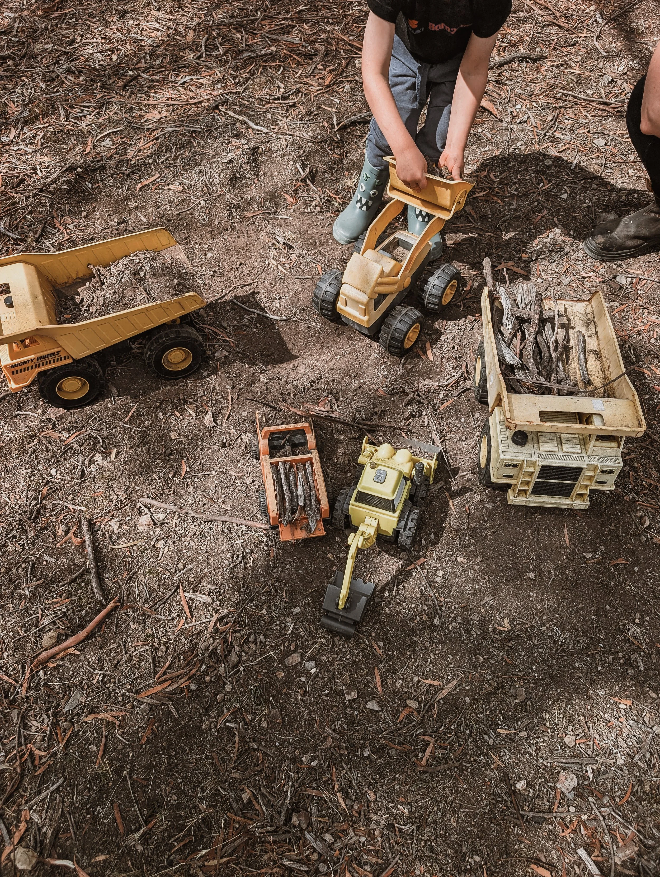 Children playing with toy construction vehicles on the ground outdoors, digging and moving dirt.