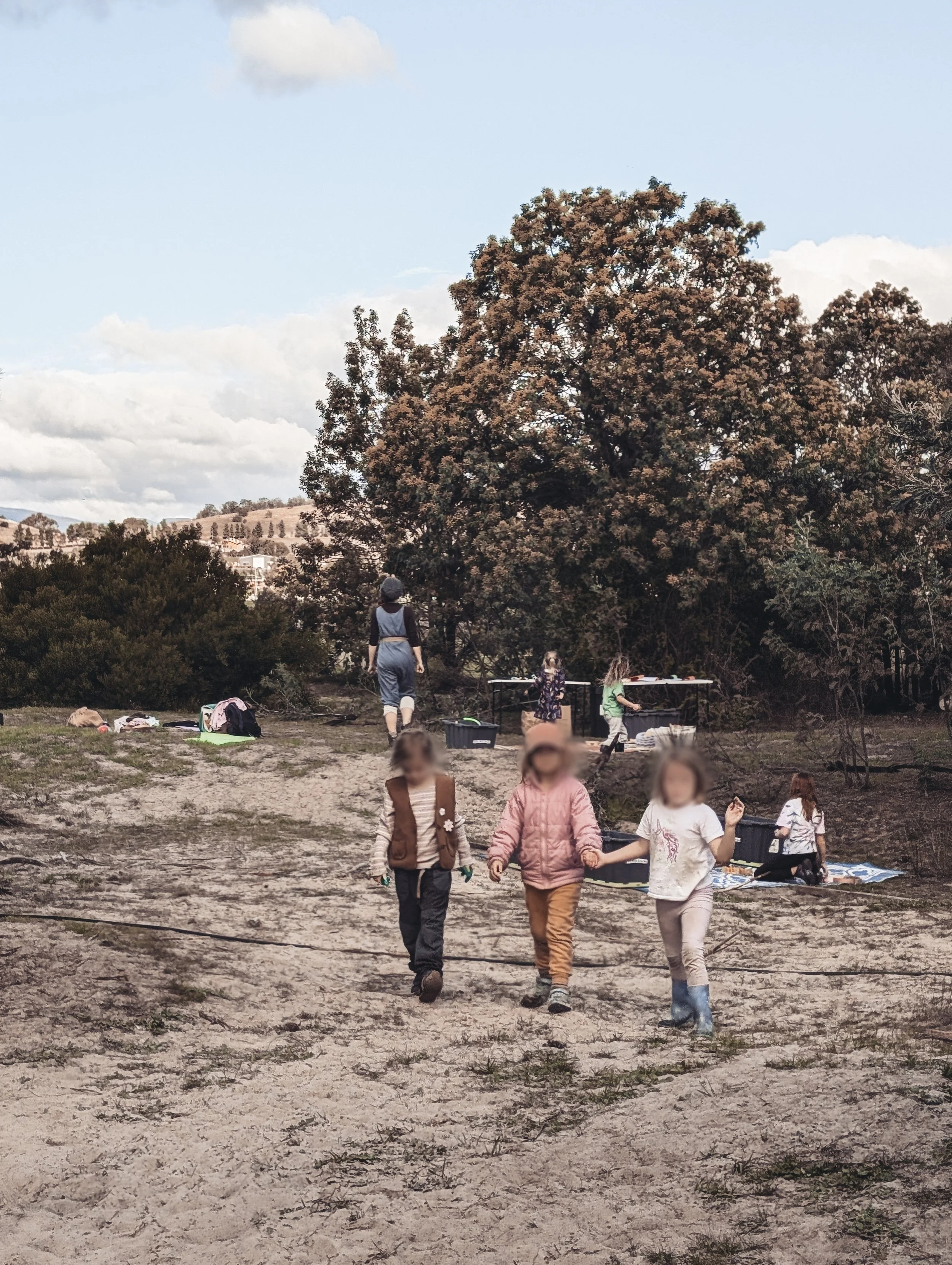 Children walking on a sandy outdoor area with greenery and trees in the background, under a partly cloudy sky.