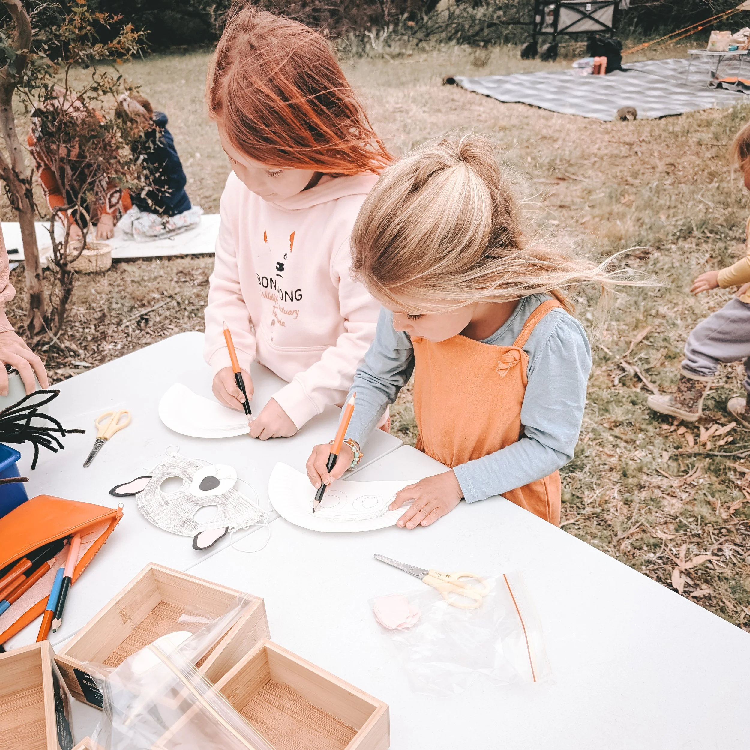 Two young girls are drawing on paper plates at a table outdoors, surrounded by craft supplies, with other children in the background near a tree and grassy area.