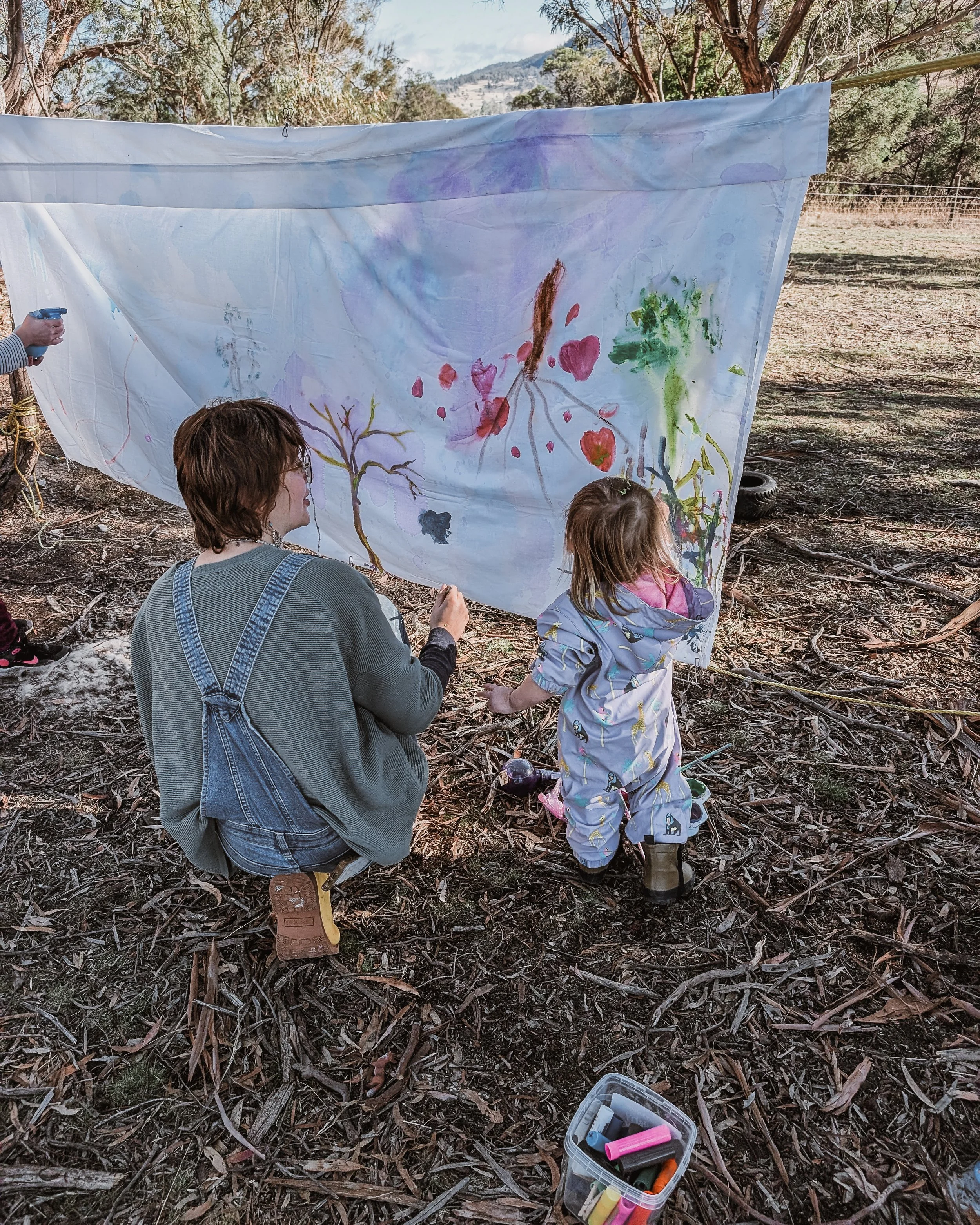 A young woman with short hair and a small child painting together on a hanging white sheet