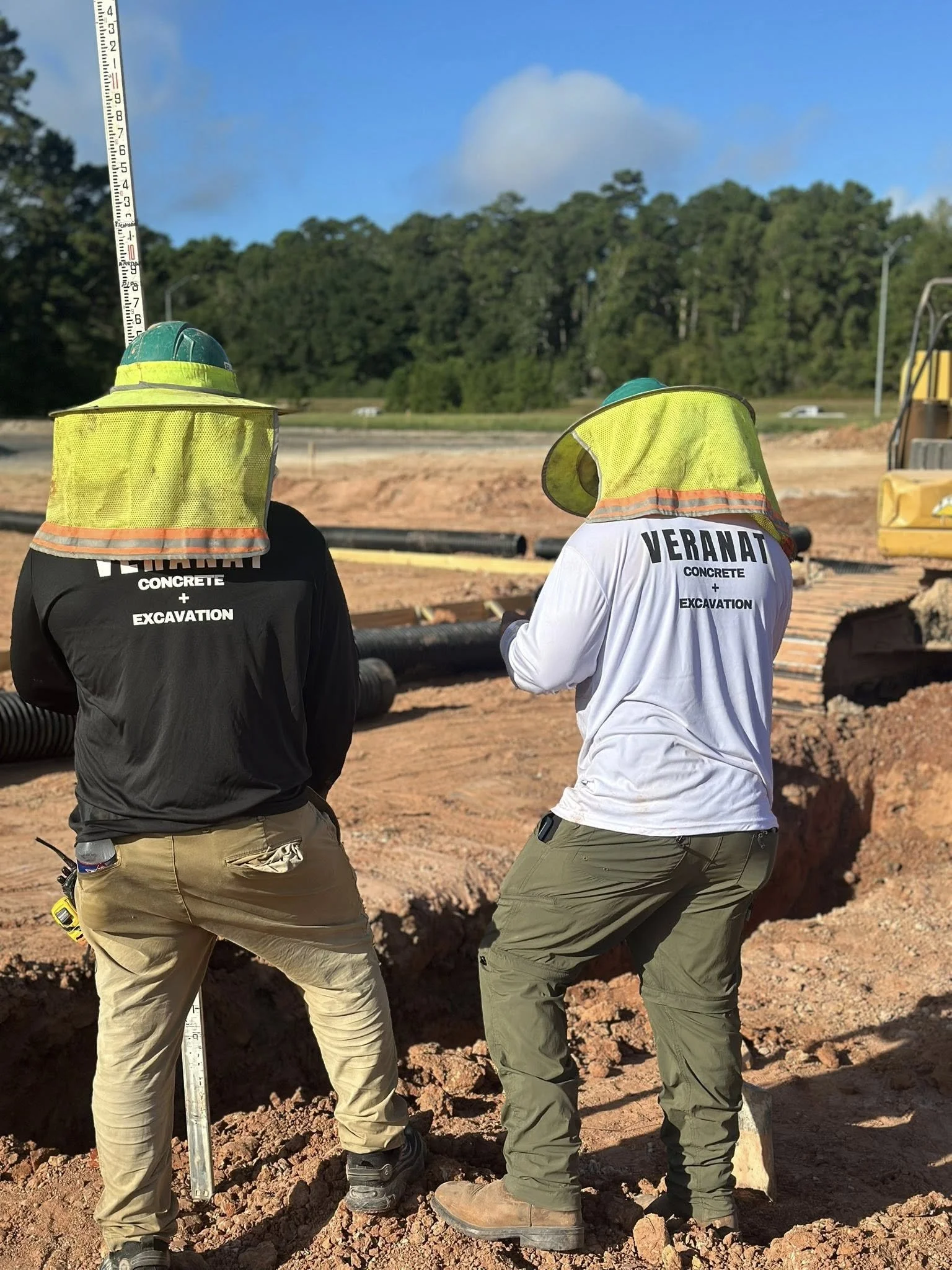 Two construction workers wearing high-visibility safety vests and helmets at a construction site, standing next to a large hole in the ground with hoses and equipment around them, and a backdrop of trees and a partly cloudy sky.