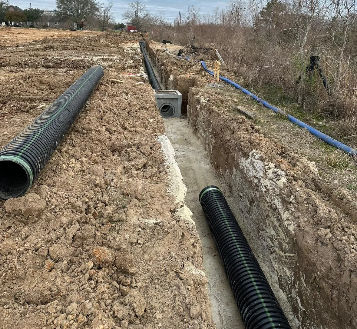 Construction site with large black corrugated pipes installed in a long, narrow trench on dirt ground, with some blue pipes seen in the background, and leafless trees to the side.