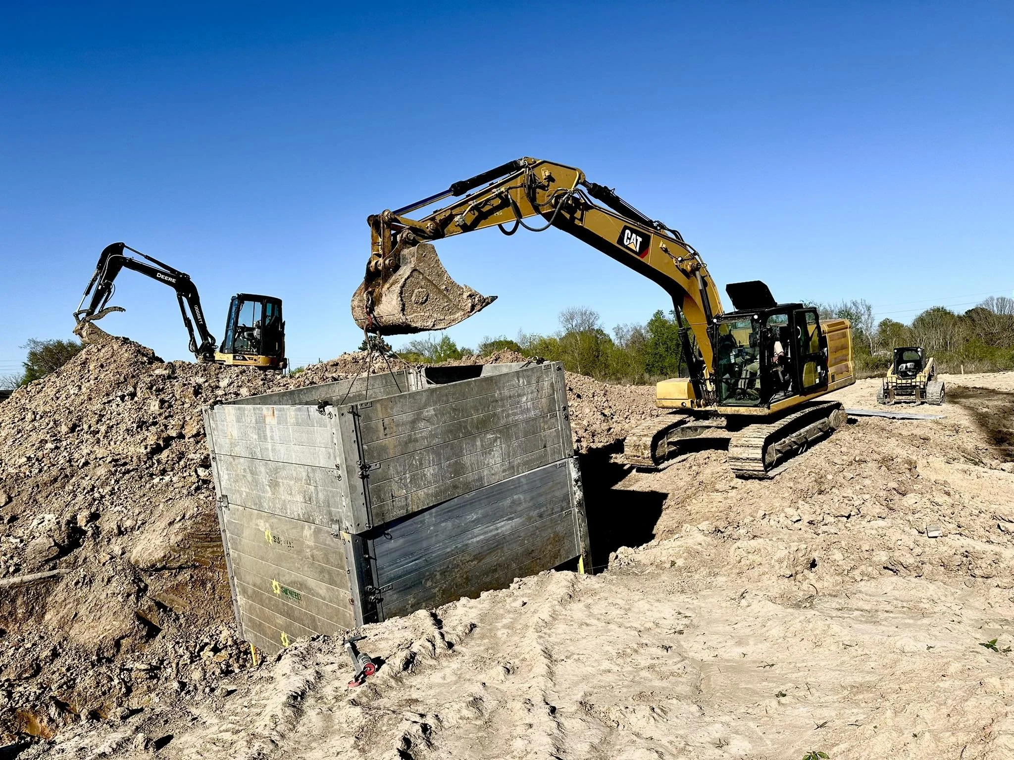 Construction site with two excavators digging and working on soil, one excavator lifting a bucket of dirt into a large metal container, and the other moving earth, under a clear blue sky.