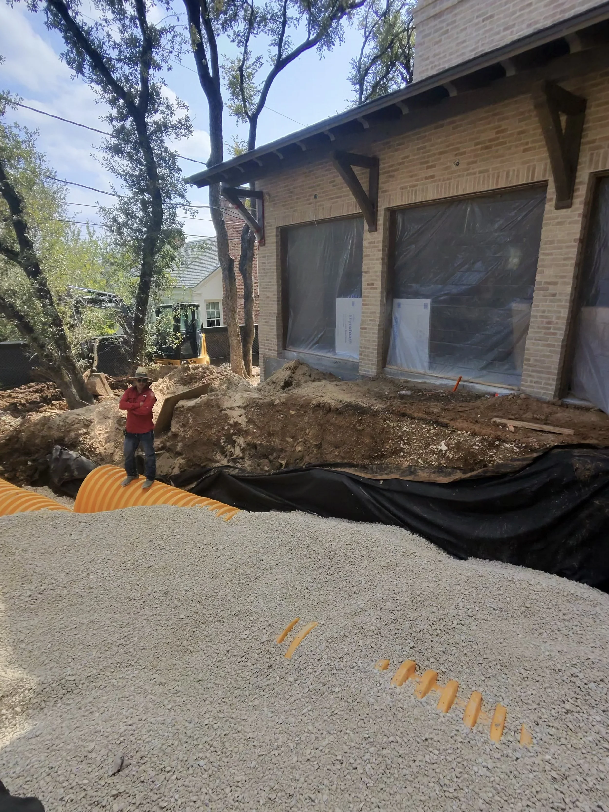 Construction site in front of a brick house, with large orange pipes, gravel, dirt, and black liner, and a worker in red shirt and jeans wearing a hat.