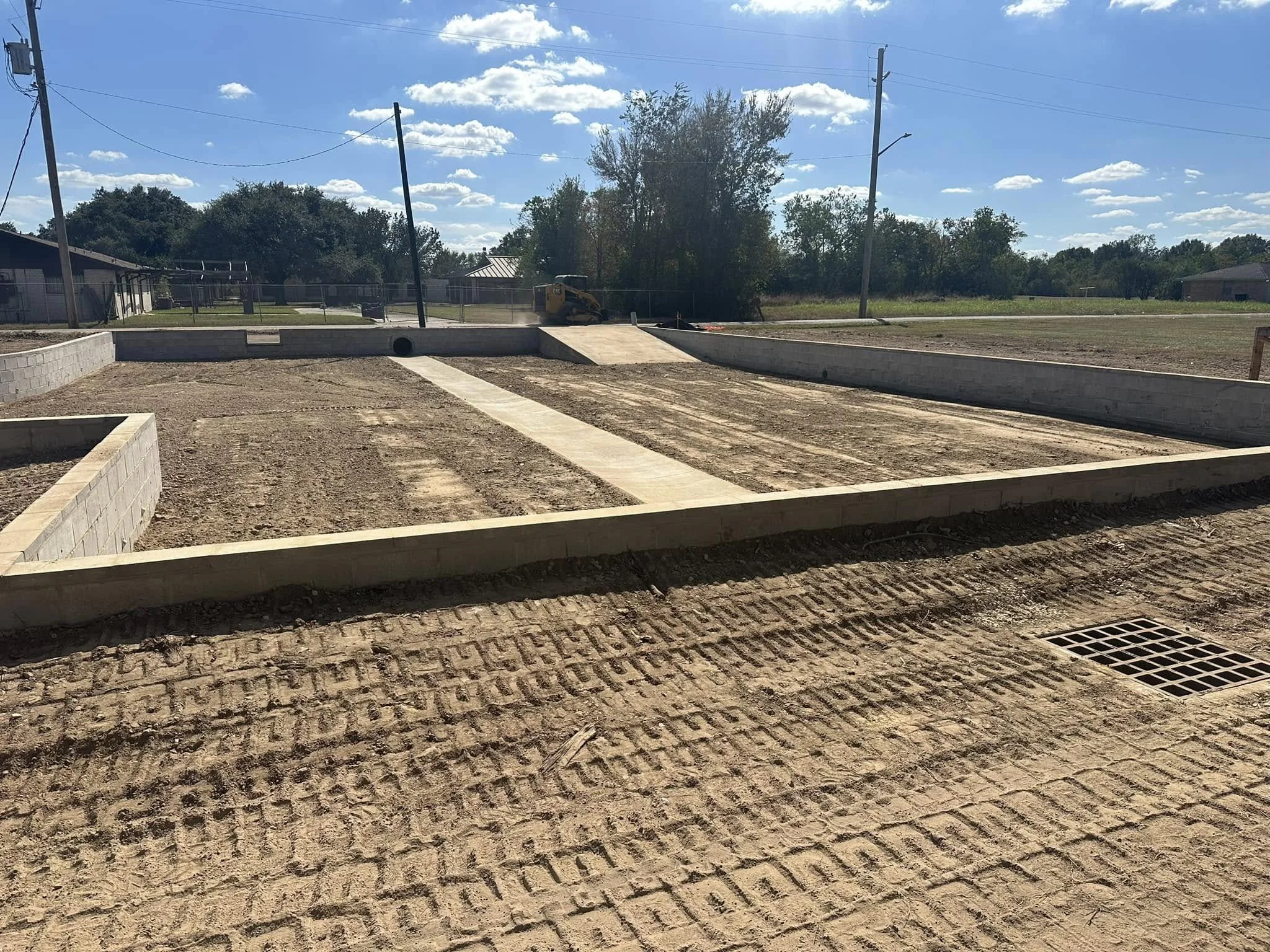Construction site with concrete foundation, dirt ground, a drainage grate, and a blue sky with clouds.