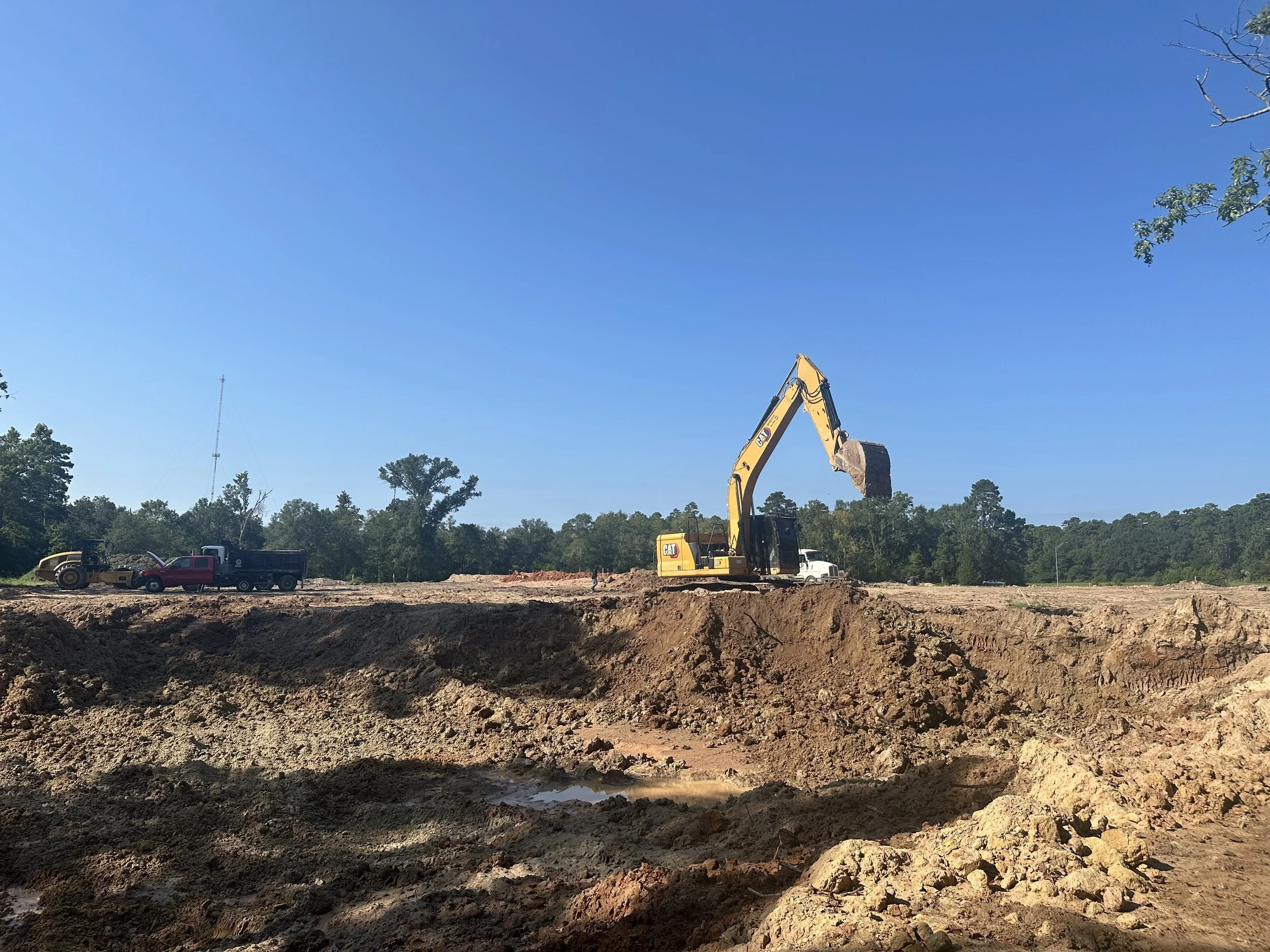 A construction site with a yellow excavator digging into the earth, accompanied by a red pickup truck and a yellow bulldozer under a clear blue sky.