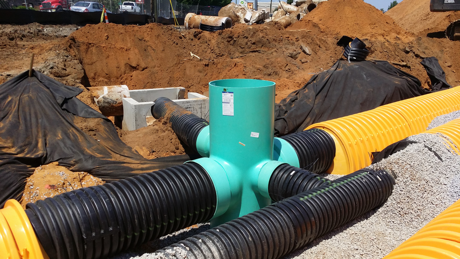 Construction site with underground piping system, including black, yellow, and turquoise pipes, surrounded by dirt and gravel.