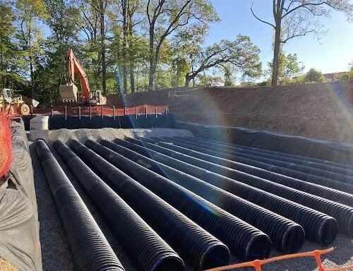 Large black corrugated pipes laid out on ground for construction, with construction equipment and trees in the background.