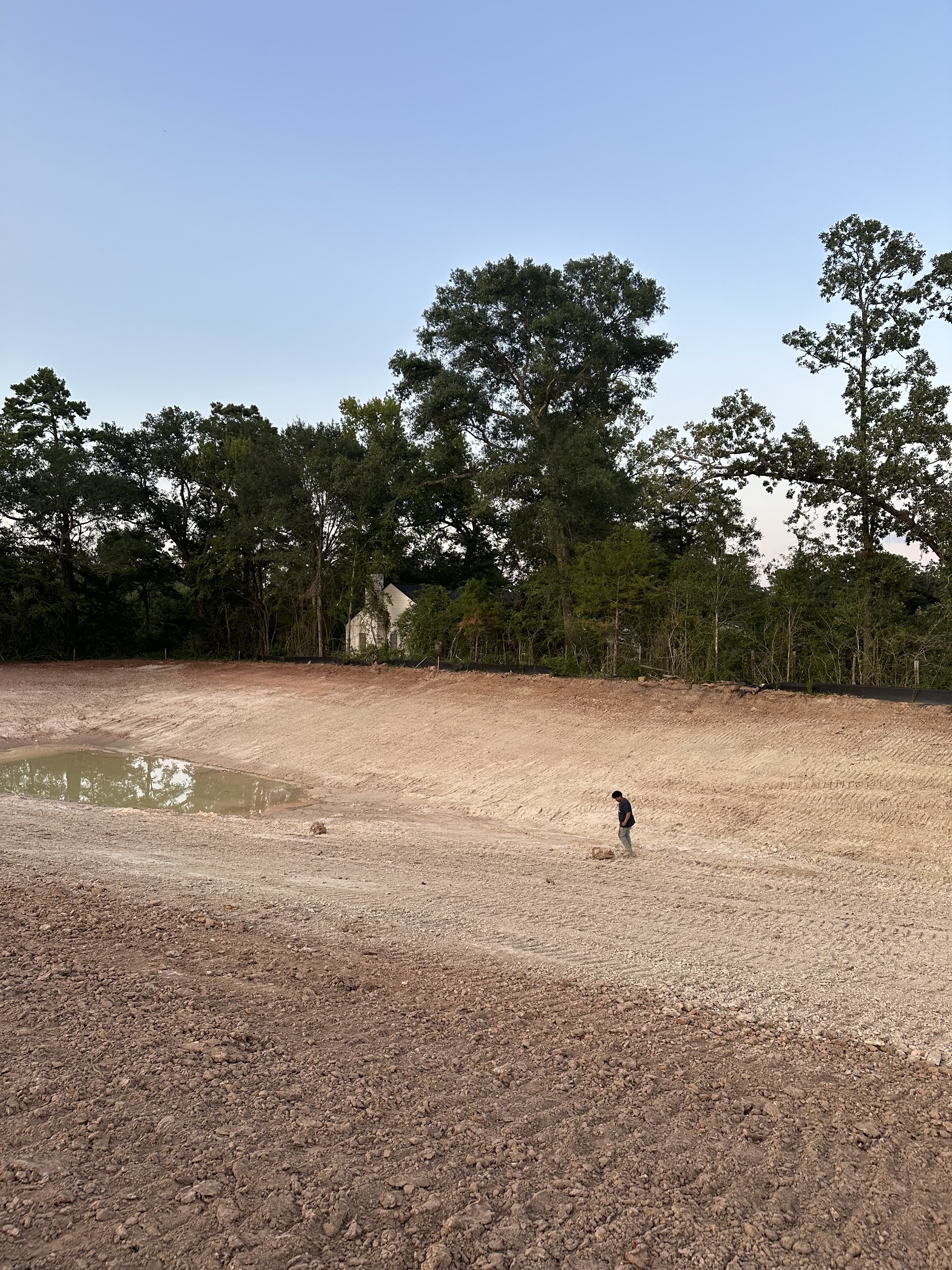 A man walking next to a small pond on a dry, dirt-covered landscape with trees in the background.