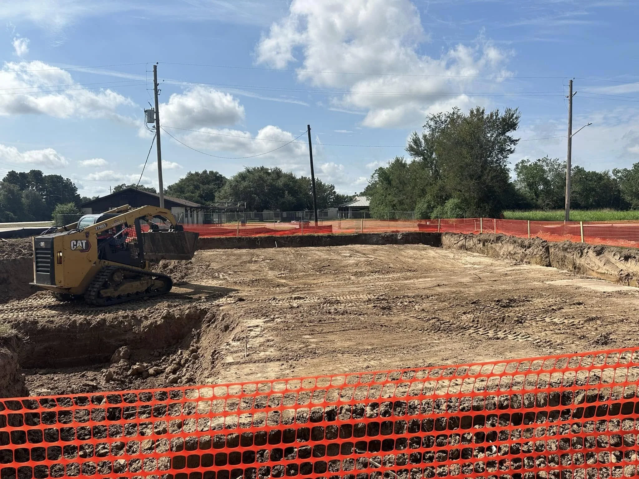 Construction site with a small tracked excavator behind orange safety fencing, with a partly dug area, utility poles, and trees in the background under a partly cloudy sky.
