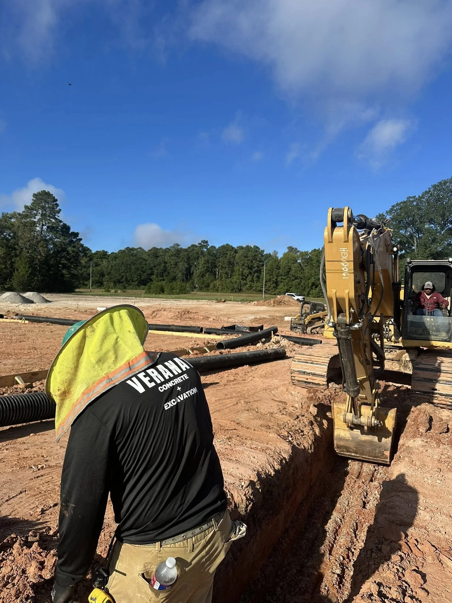 Construction worker wearing a yellow safety helmet and black shirt with 'Veraha Concrete + Excavation' printed on back. The worker is standing near an open trench at a construction site with an excavator and dirt in the background, under a blue sky w