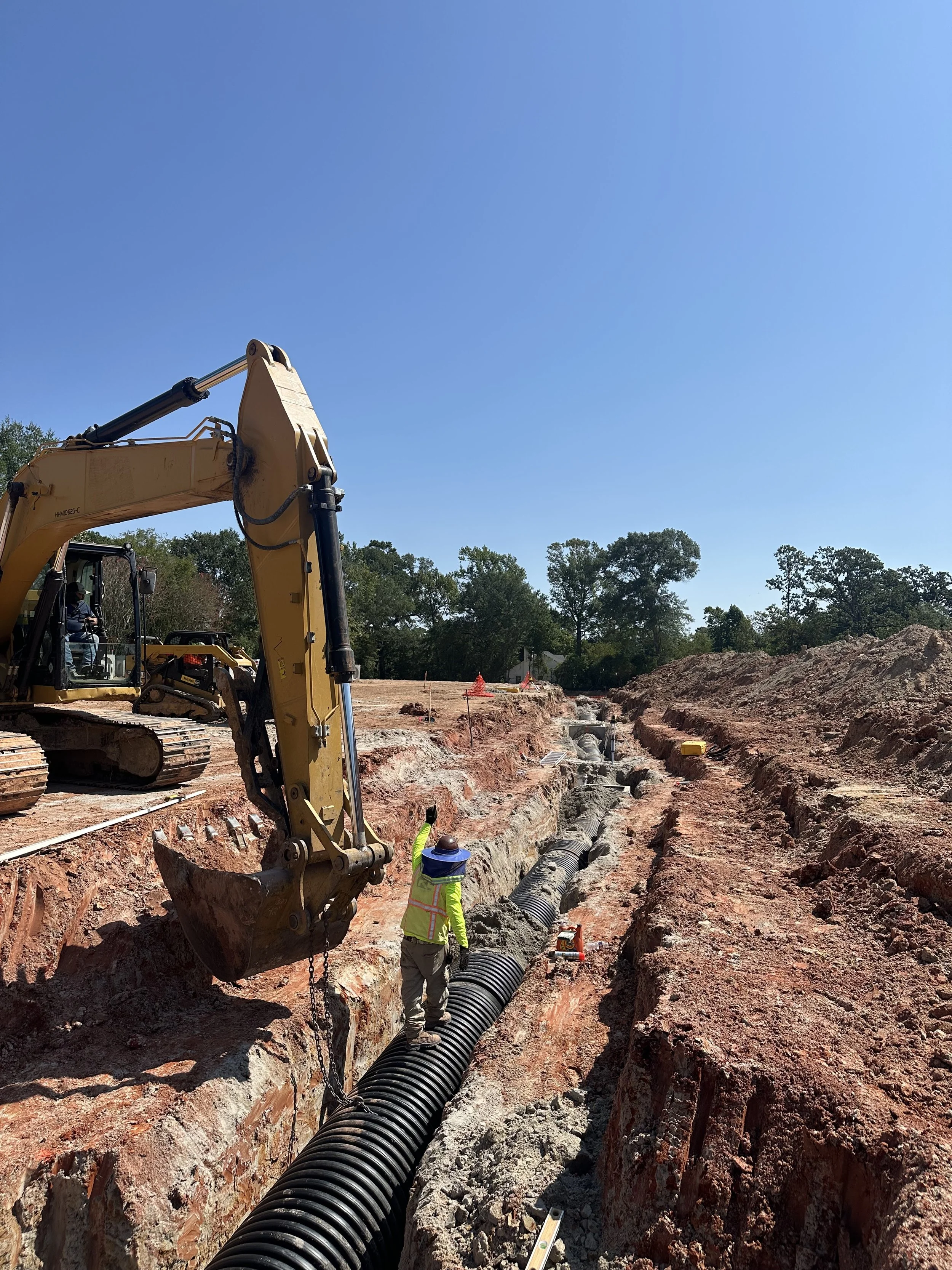 Construction site with a large excavator working on underground pipe installation, worker in a yellow safety vest walking on black corrugated pipe, under a clear blue sky.