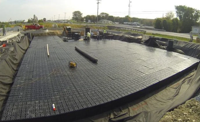 Construction workers installing a black plastic foam drainage system on a building foundation, with a large water-resistant tarp around the perimeter.