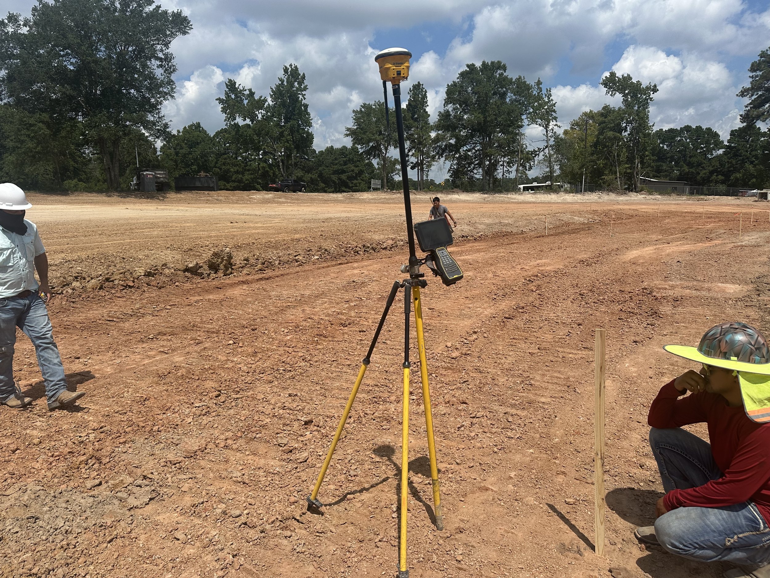 Construction workers on a dirt site using surveying equipment, with one worker squatting and another standing, in a partially cleared outdoor area with trees and cloudy sky in the background.