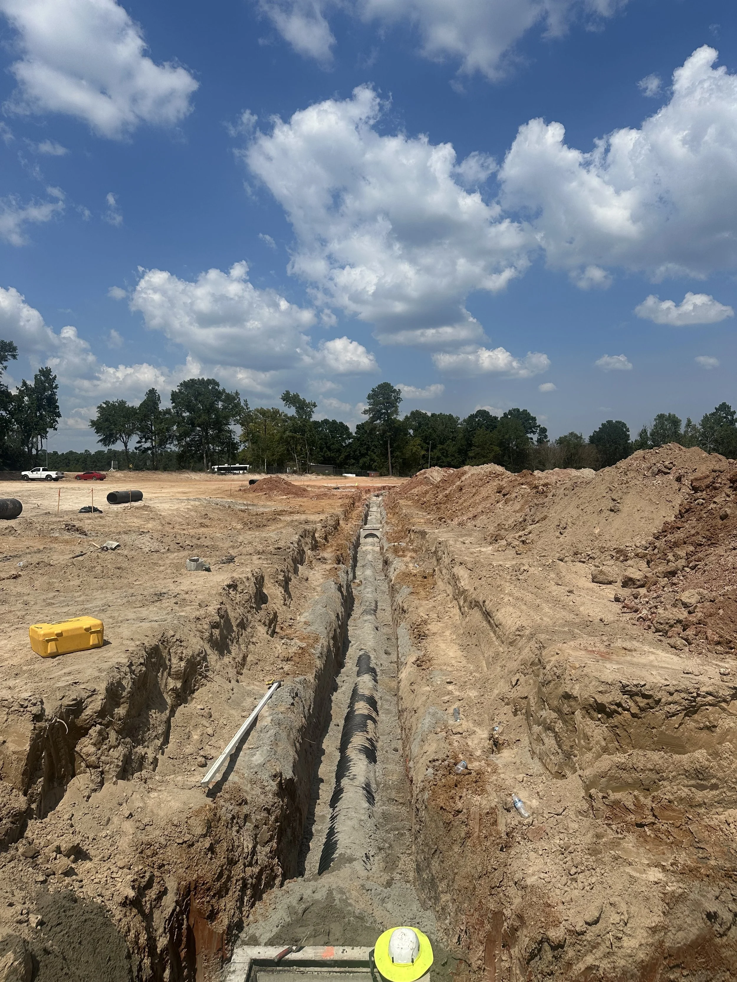 Construction site with a deep trench for pipe installation, scattered tools, and a worker helmet at the foreground, with a partly cloudy blue sky and trees in the background.