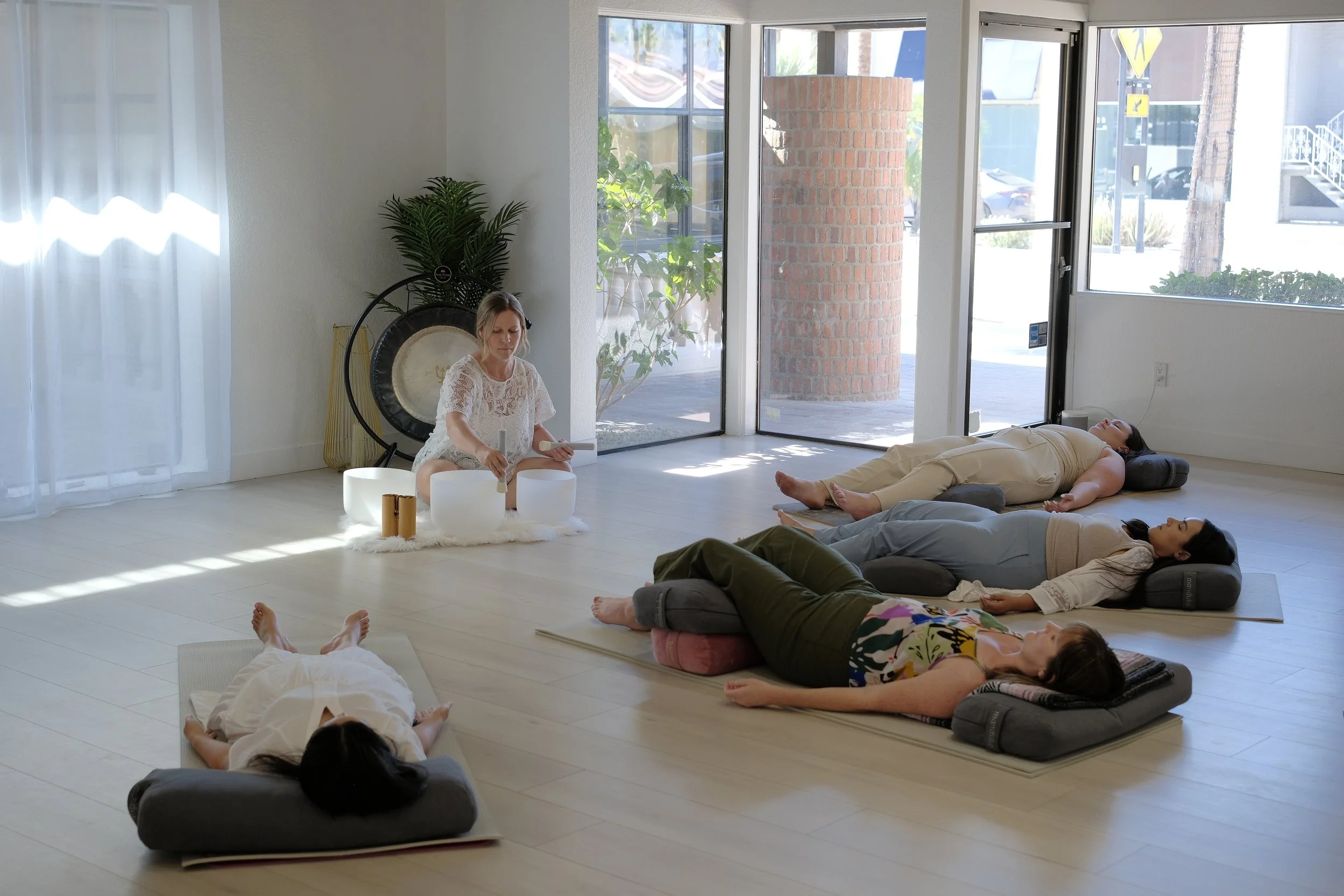 People practicing sound healing or meditation in a bright, airy room with large windows, cushion seats, and a musical gong.