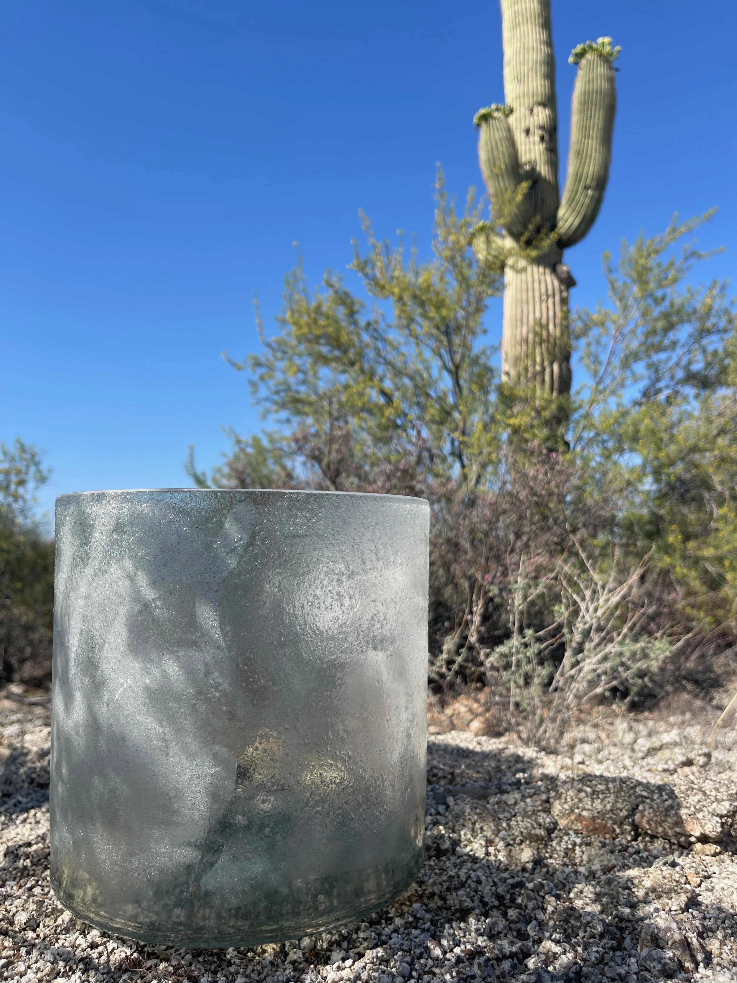 Close-up of a frosted glass candle holder on gravel ground with a cactus and desert plants in the background under a clear blue sky.