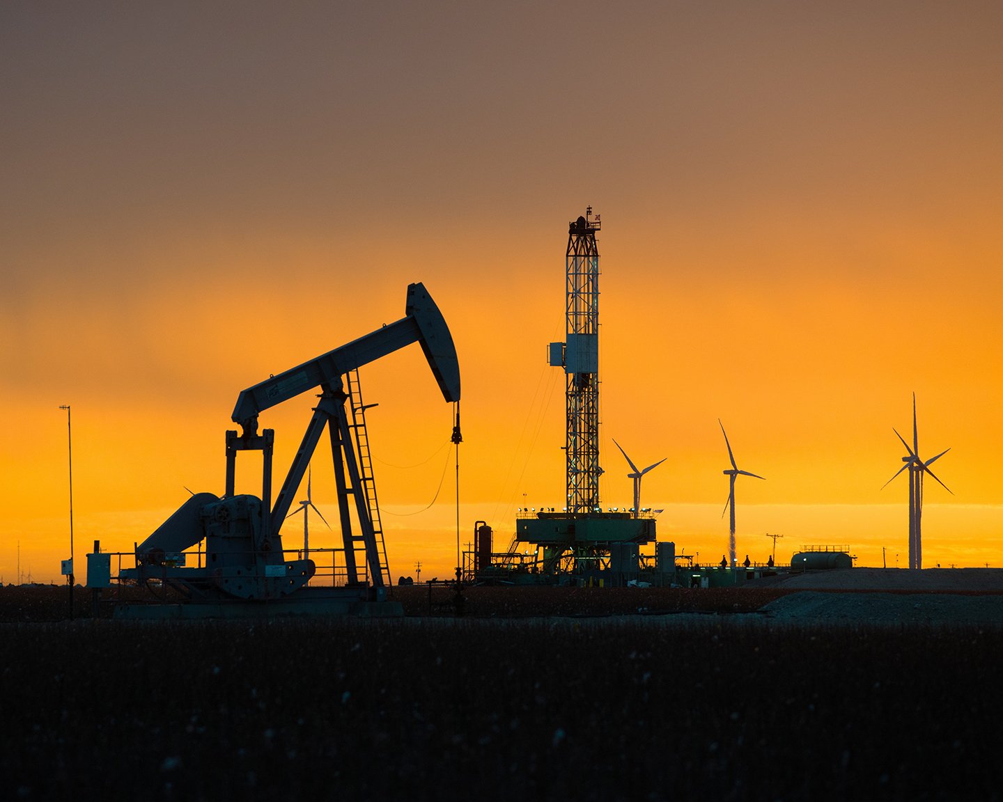 A pumpjack and drilling rig stand among wind turbines at sunset.