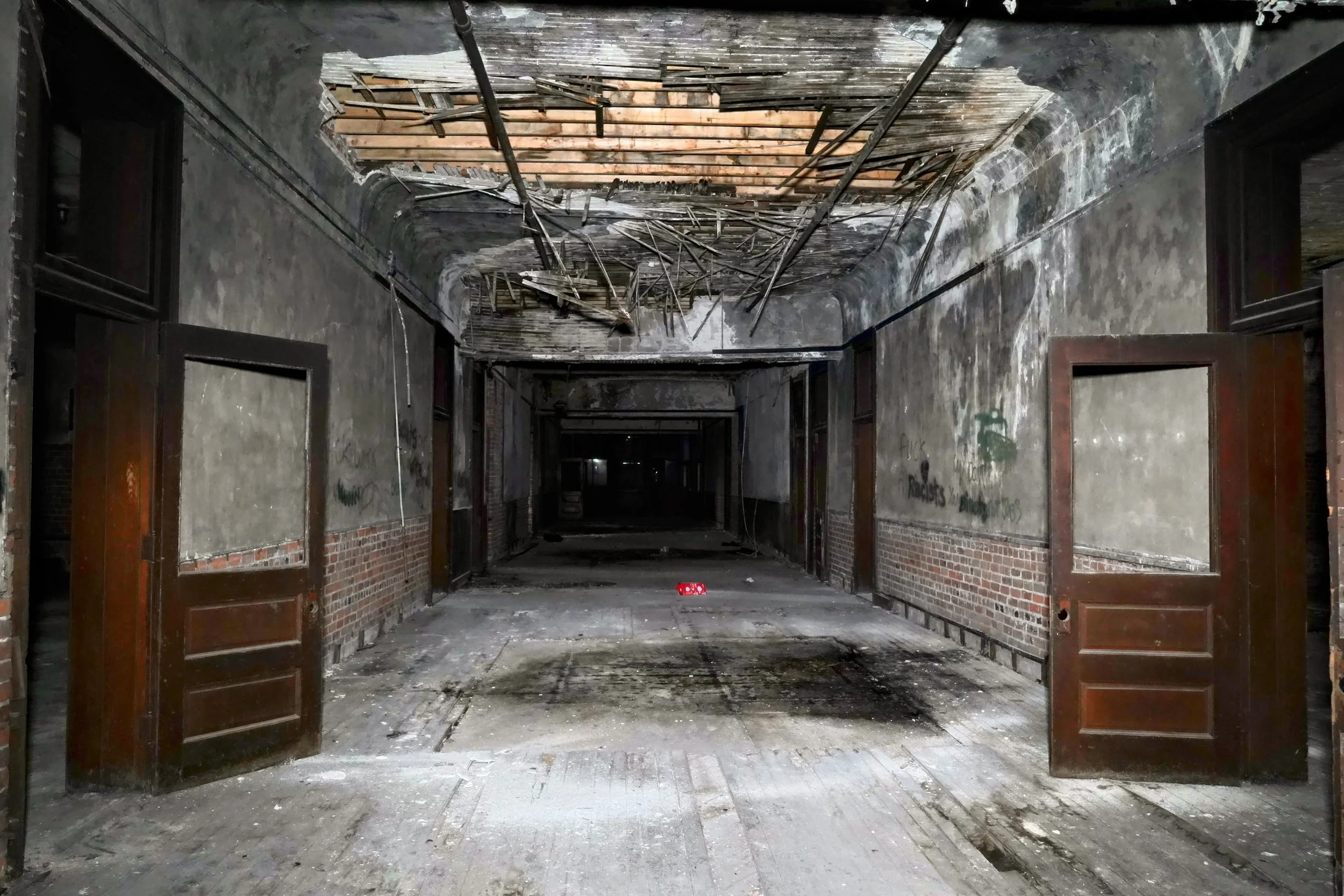 Dilapidated hallway with broken ceiling, peeling paint, and open wooden doors.