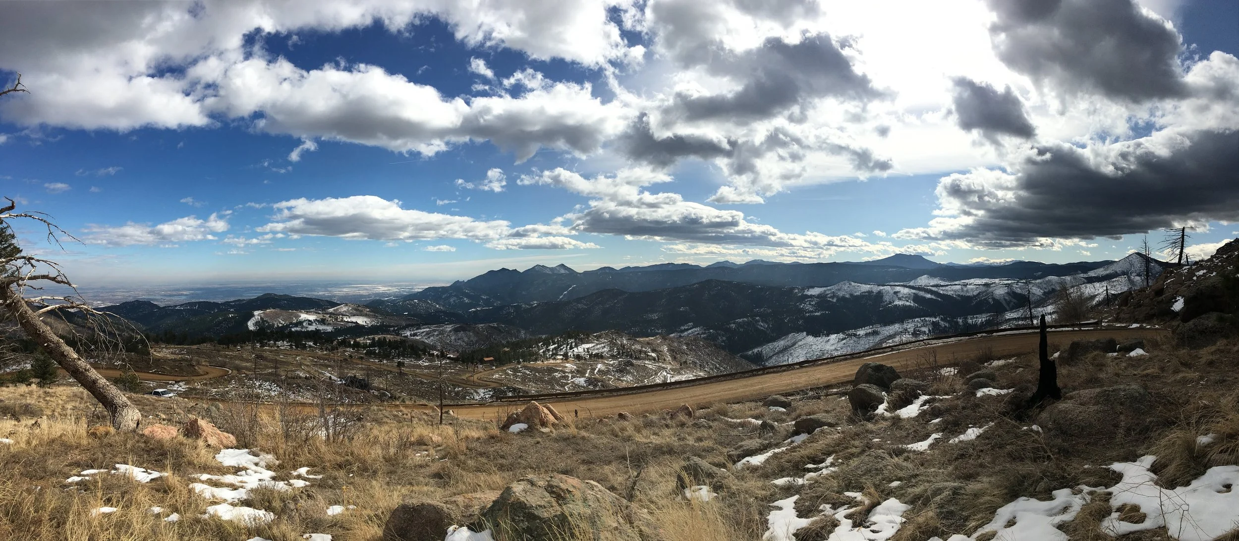 Scenic mountain landscape with snow patches, grassy foreground, winding dirt road, and dark cloudy sky.