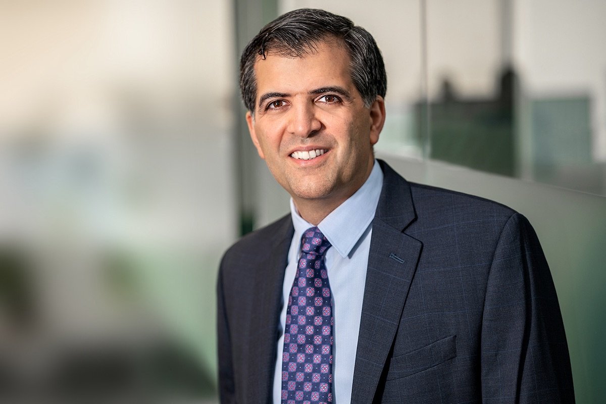 Corporate headshot of smiling man standing in an office with defocused office background.