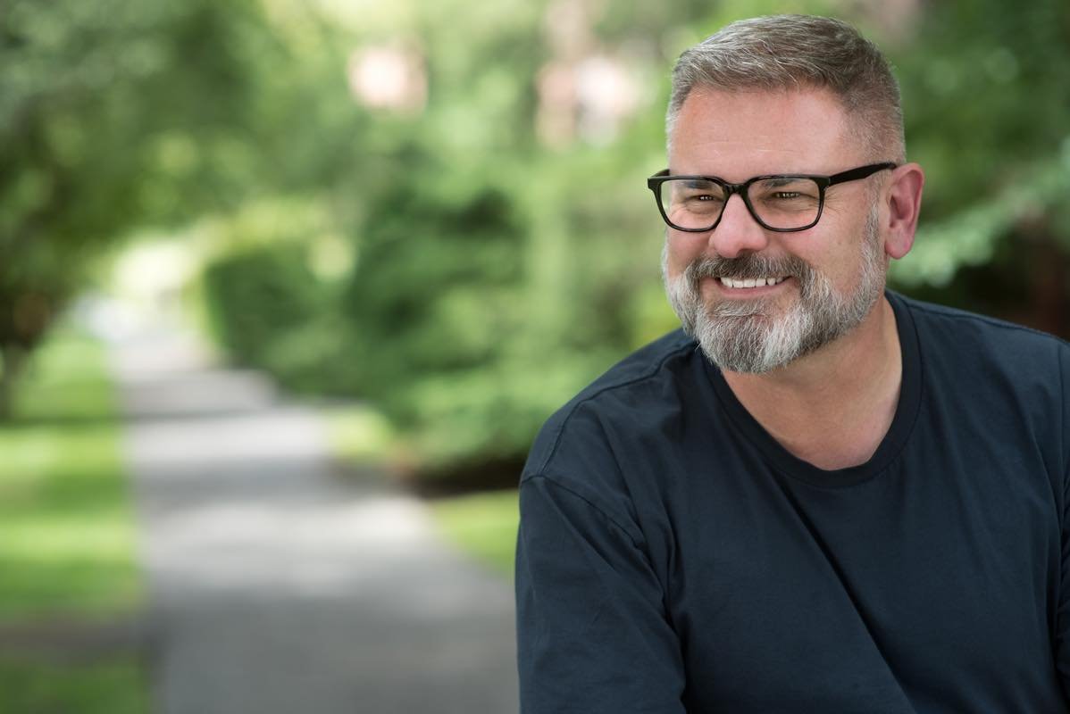 Branding portrait of man outside looking to left, greenery in background.