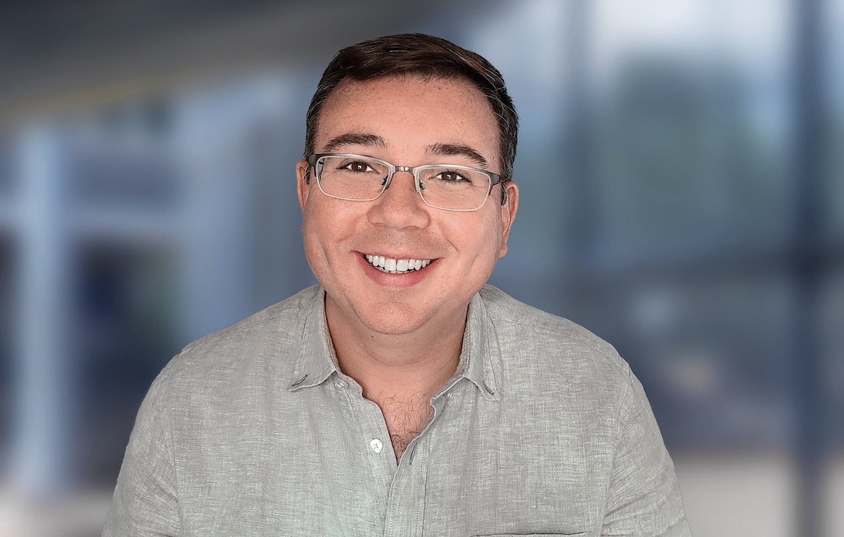 Casual headshot of a man in an open-collar gray shirt with an out-of-focus foyer background.