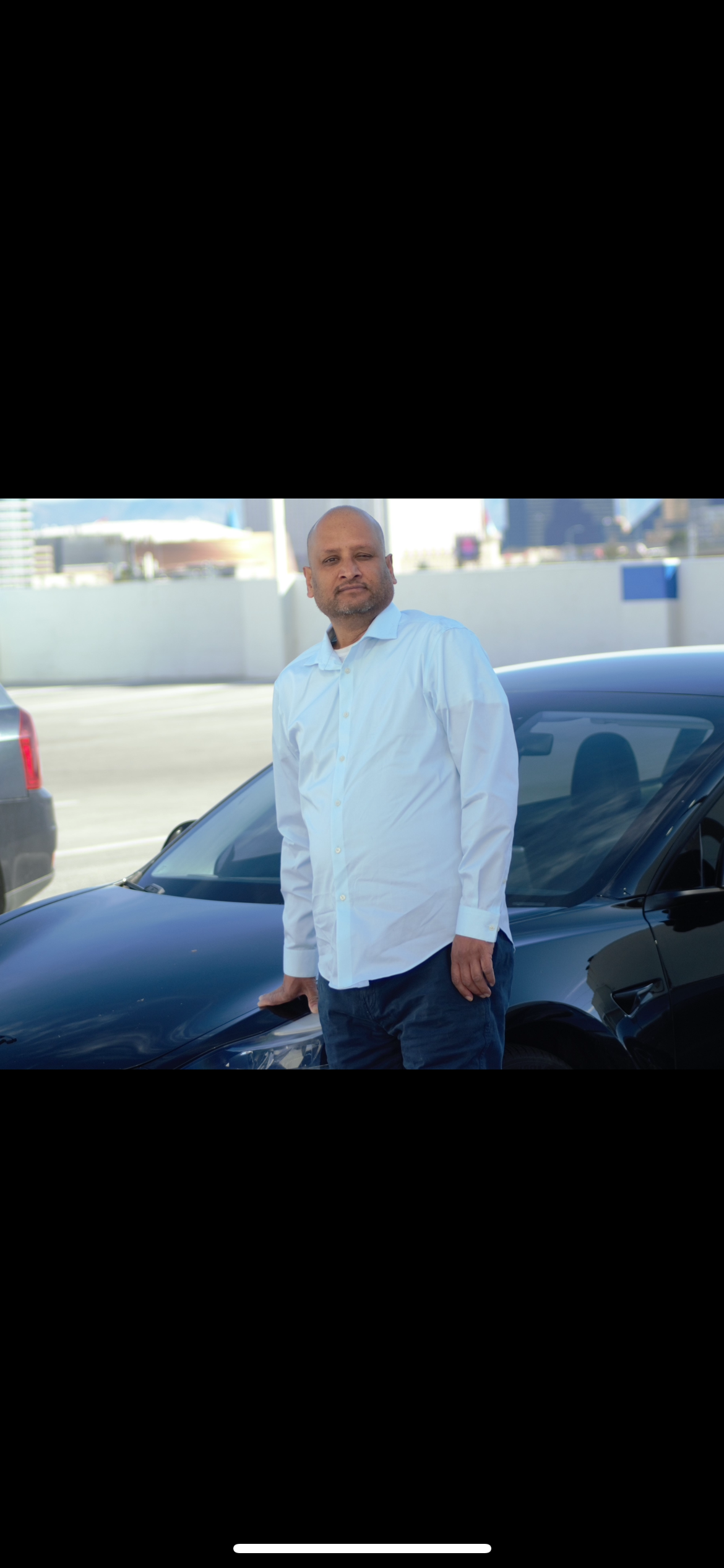 A man in a white shirt and dark pants standing beside a black sports car on a rooftop parking lot, with a city skyline in the background.