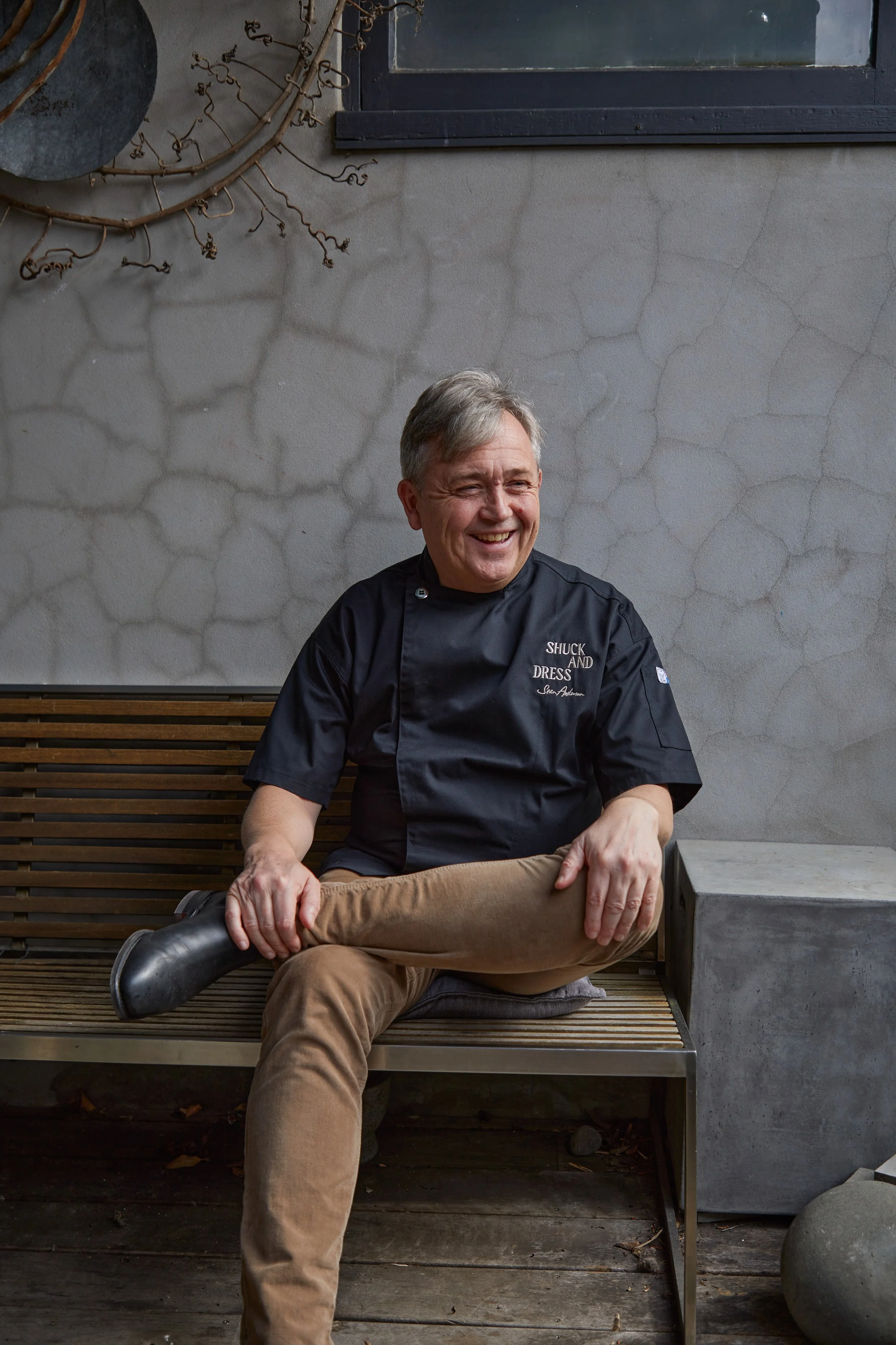 A man with gray hair is sitting on a wooden bench, smiling, and holding his left foot with his left hand. He is wearing a black chef's coat and tan pants, with a black boot on his right foot. The background features a textured gray wall and some decorative wall art.