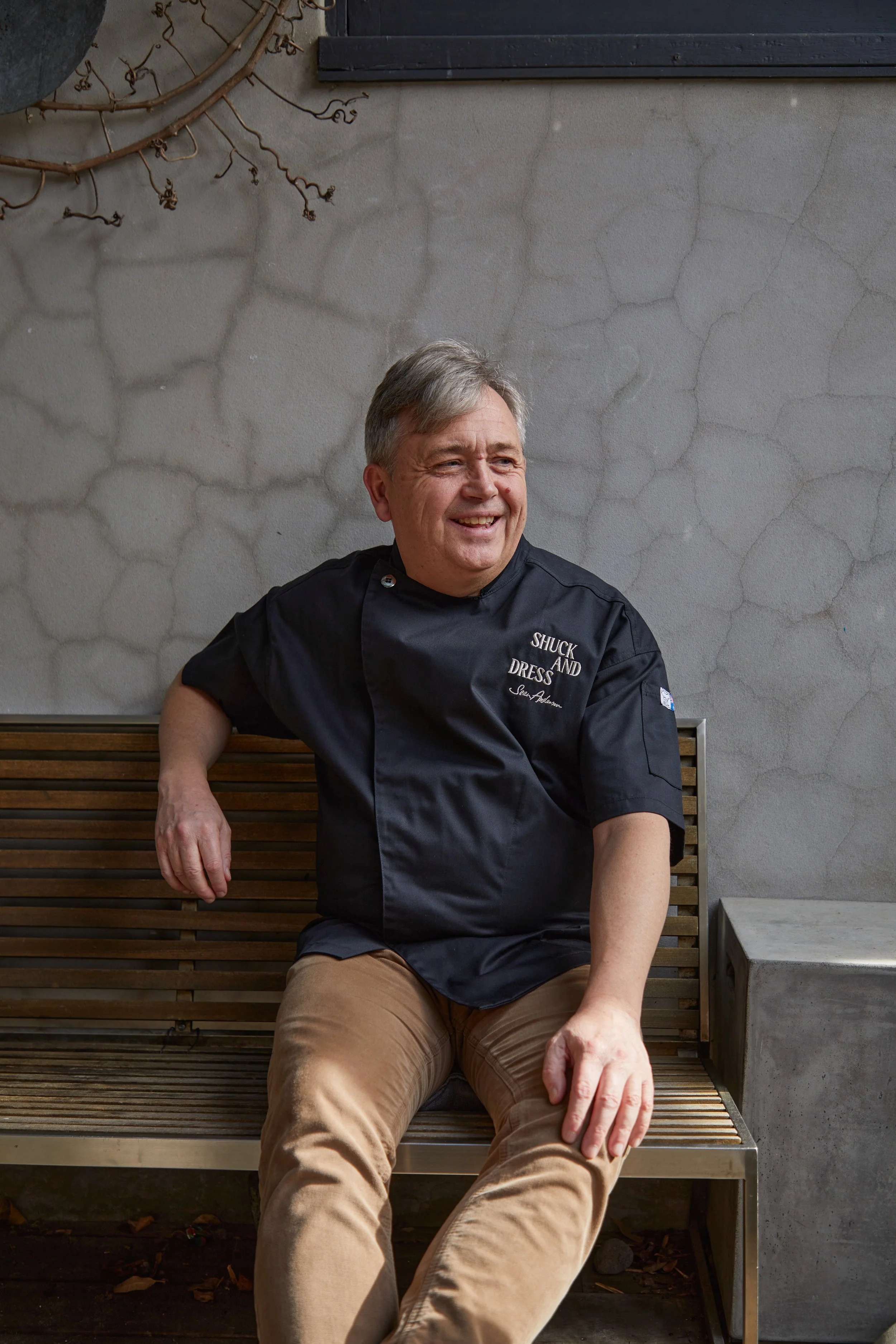A cheerful man in a black chef's jacket featuring the words 'Shuck and Dress' and other words, sitting on a wooden bench outdoors against a textured concrete wall.