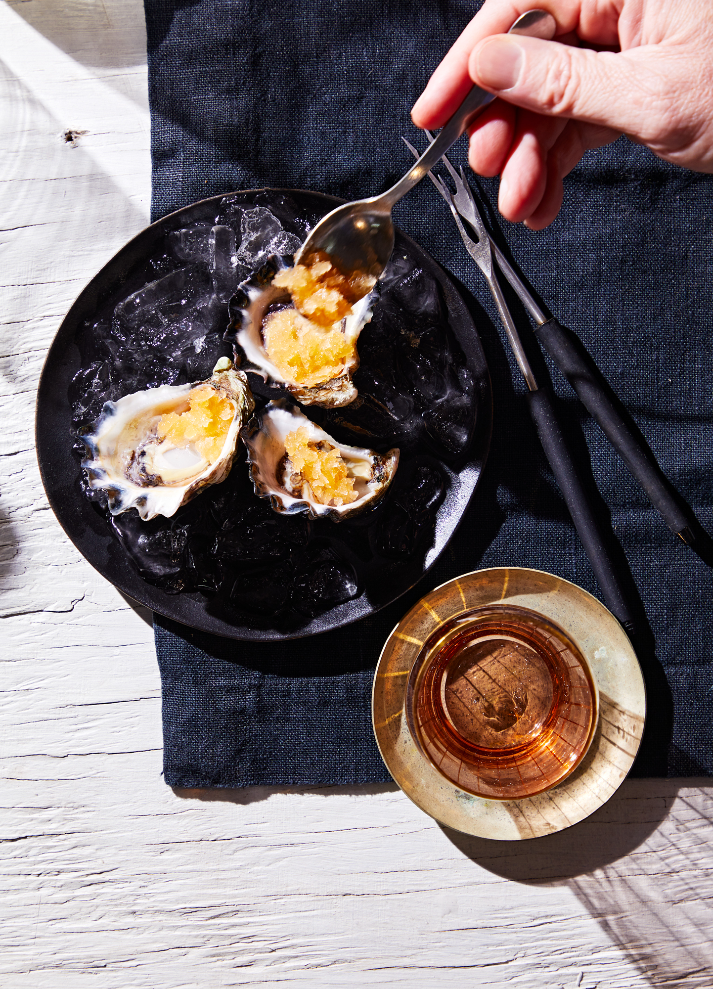 Three oysters topped with a yellow sauce on a bed of ice on a black plate, with a glass of amber-colored beverage nearby on a beige saucer, all placed on a white wooden surface with a dark cloth underneath.