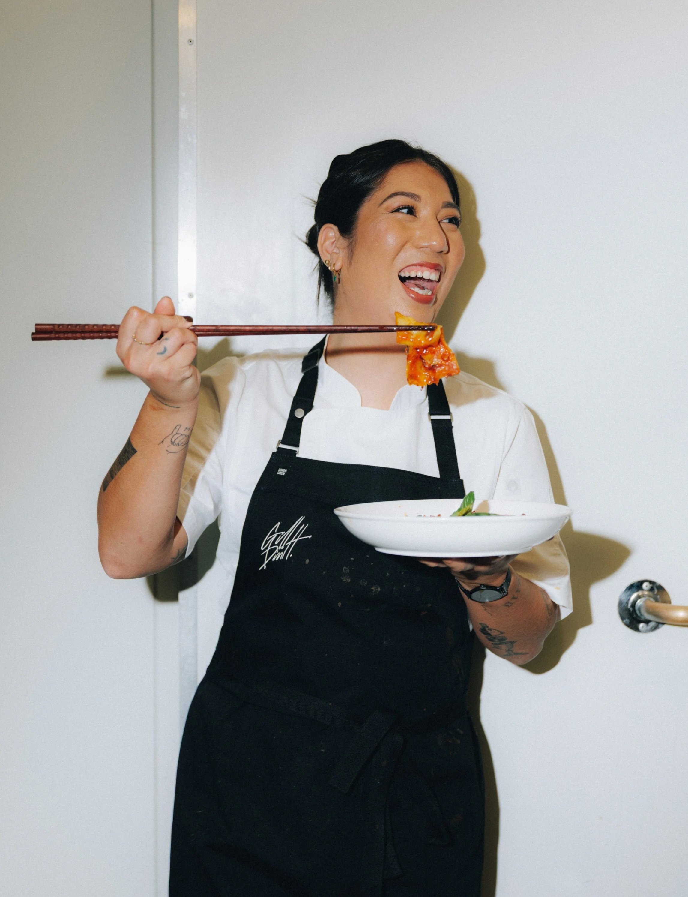 A woman in a white chef's coat and black apron holding a bowl of food and using chopsticks, smiling and looking to the side.