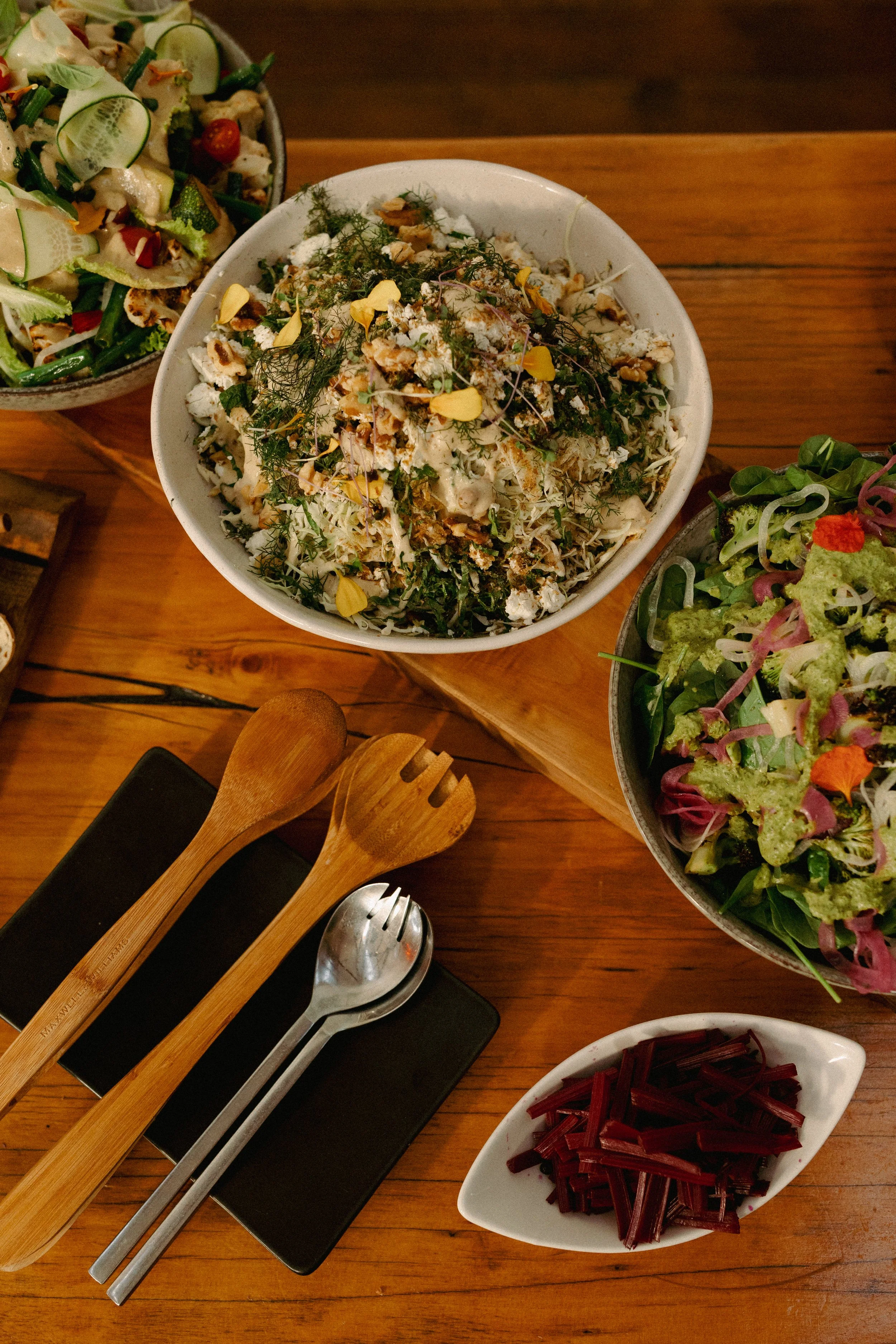 Various bowls of fresh salads on a wooden table, including one with shredded vegetables and herbs, and another with mixed greens and toppings. There is a small dish of cooked red beets, and wooden and metal salad utensils.