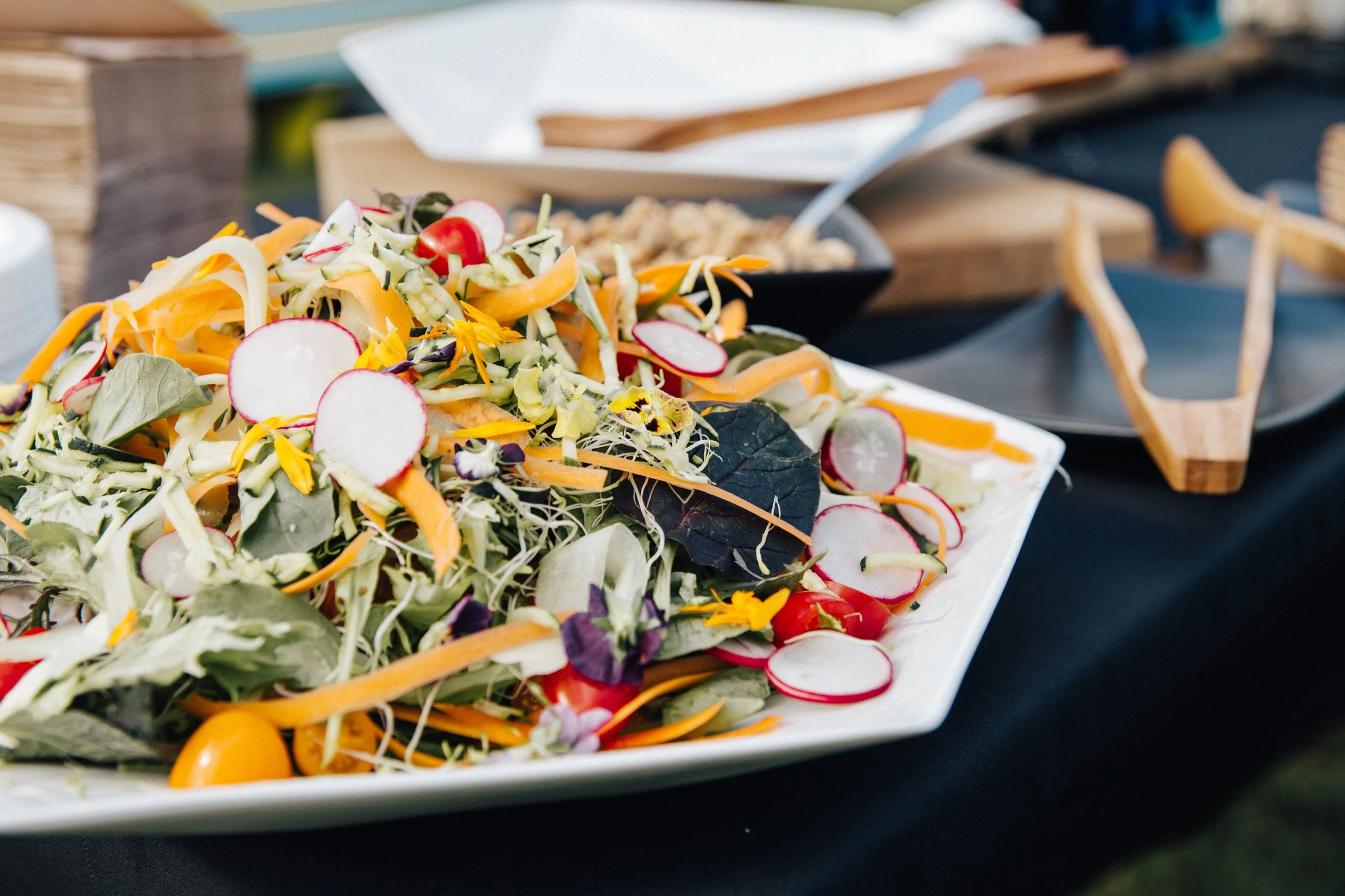 Mixed fresh salad with radishes, cherry tomatoes, shredded carrots, and various greens on a white platter at an event.