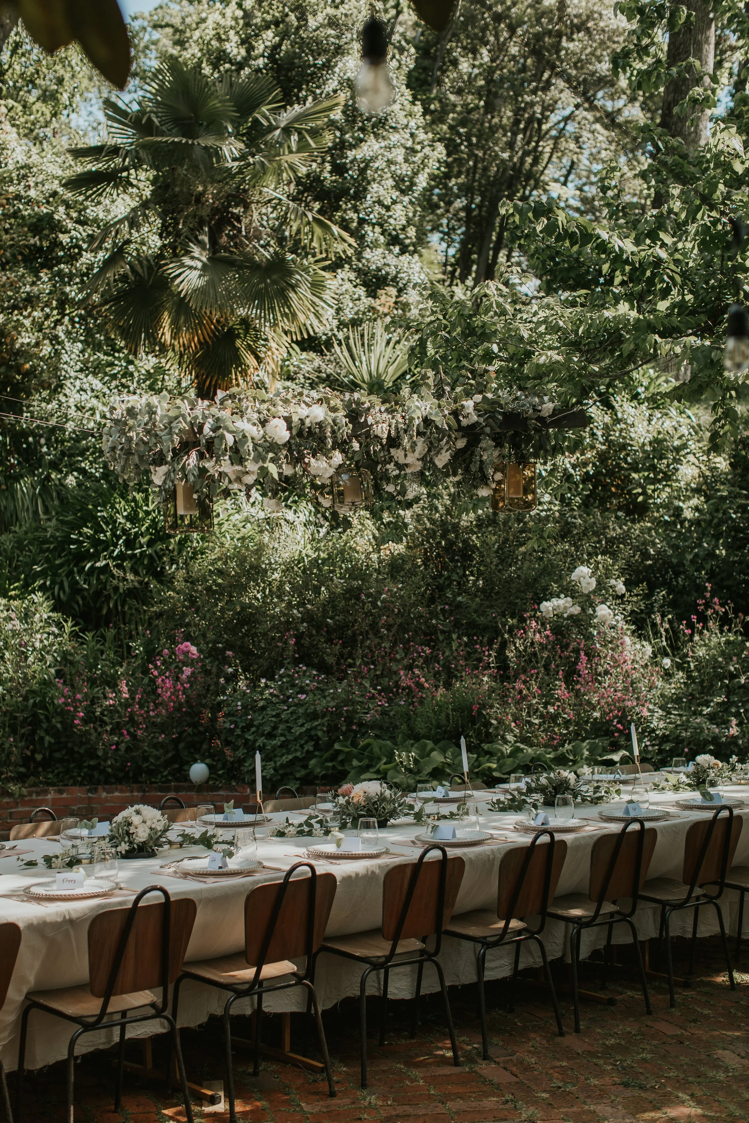 Long outdoor table decorated for a celebration, set with white plates, floral centerpieces, candles, and glassware, surrounded by lush green plants and trees in a garden setting.
