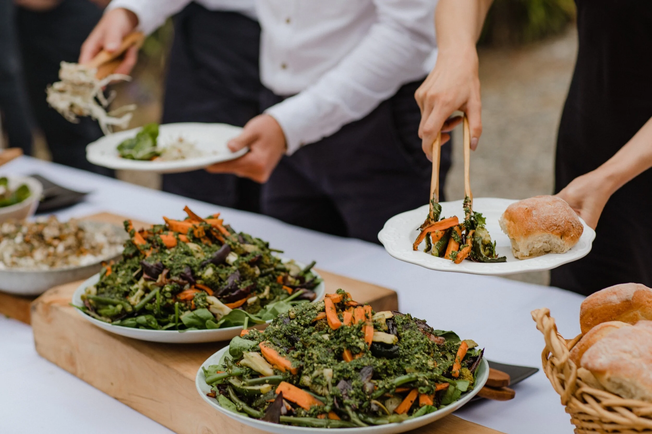 People serving themselves food at a buffet table with various salads and bread rolls. Event catering image.