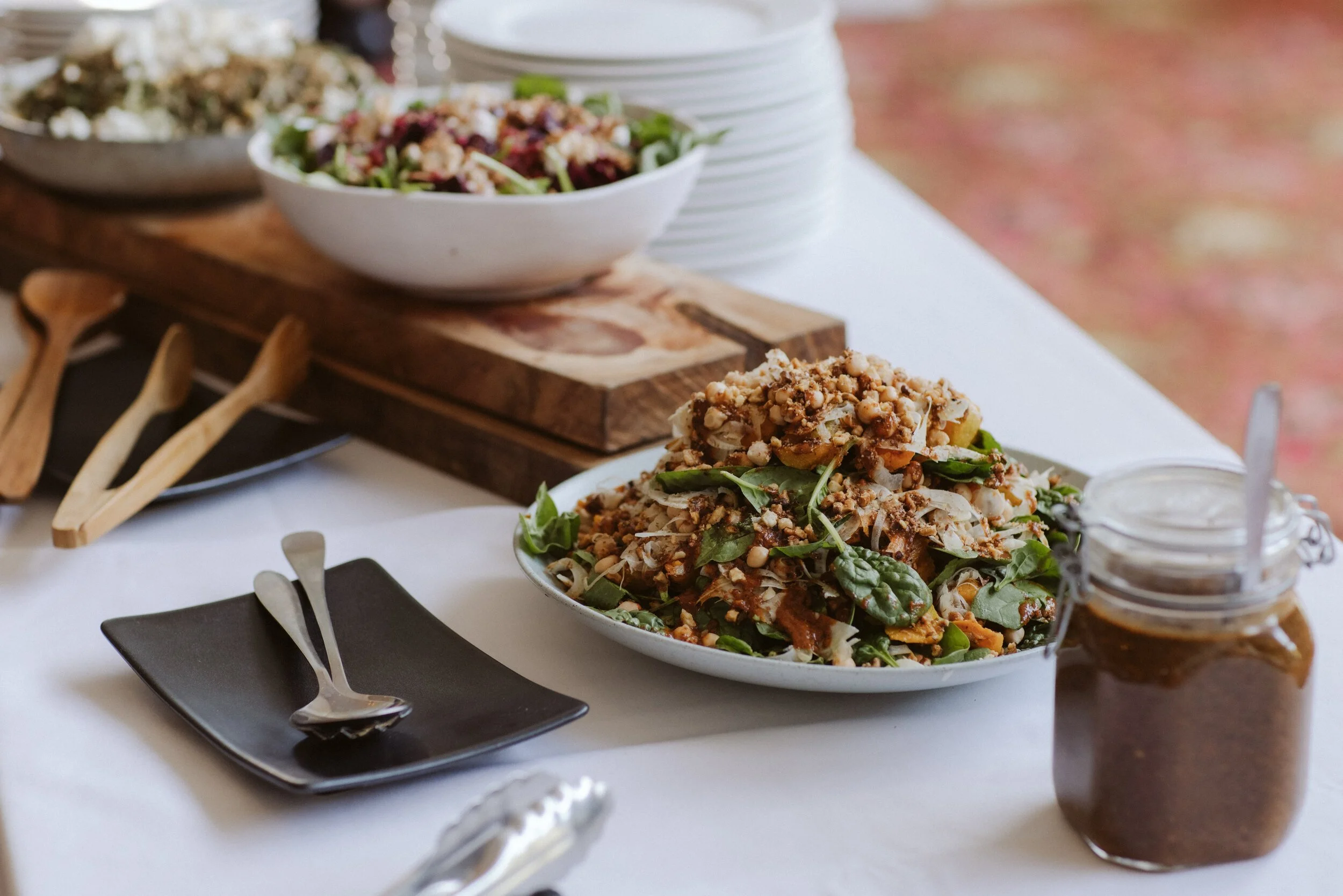 A white platter of salad topped with nuts and greens on a table, with a jar of dressing nearby and more salad bowls in the background.