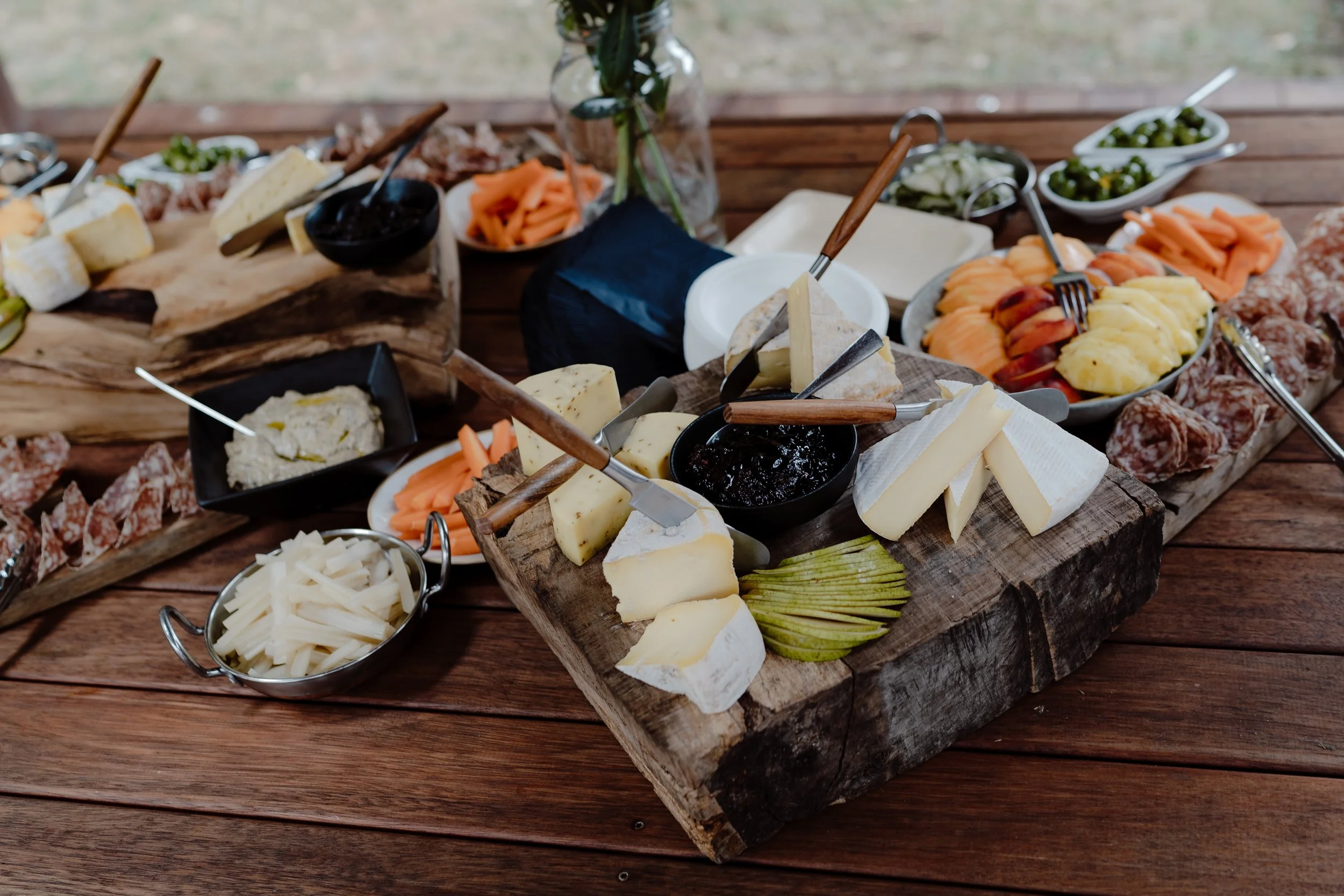 Variety of cheeses, sliced meats, and vegetables on a rustic wooden table, part of a cheese platter or charcuterie board.