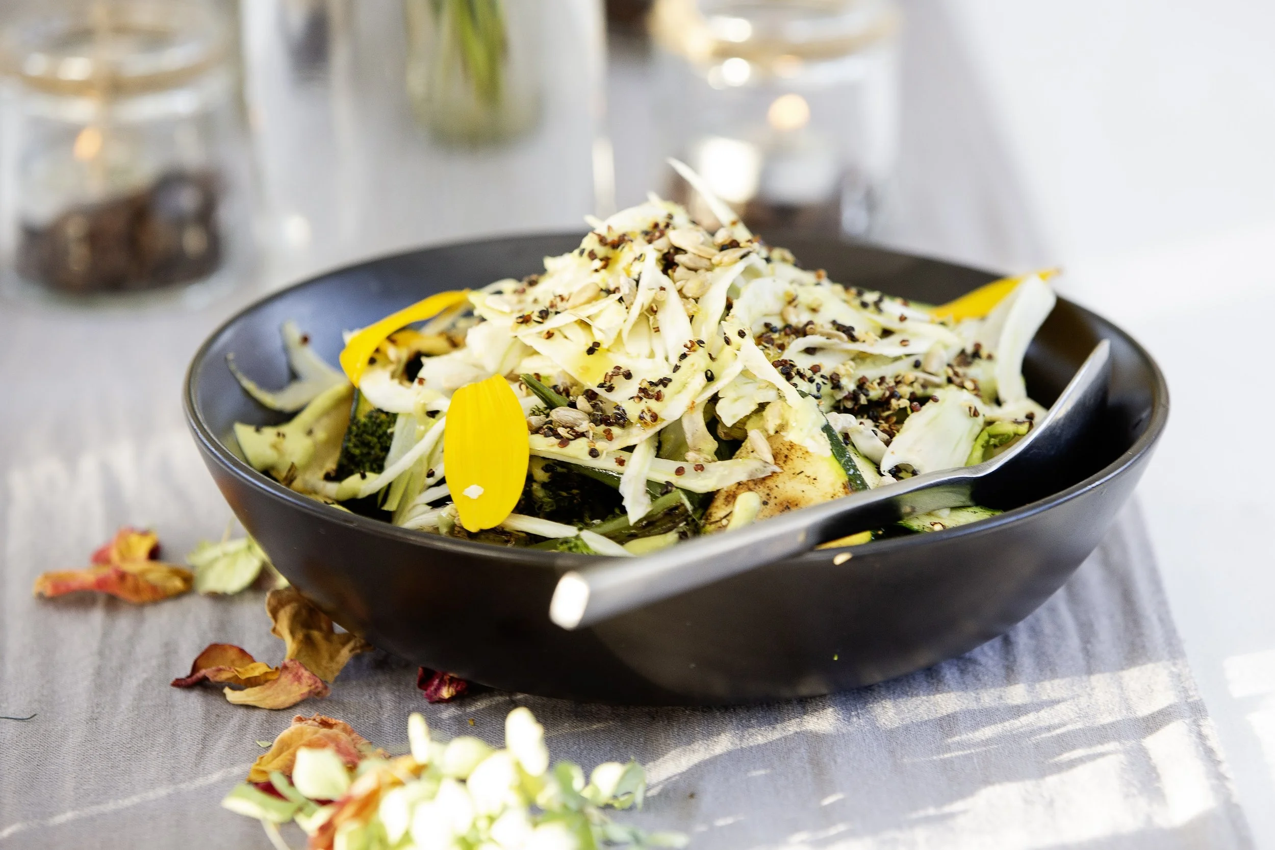 A black bowl of salad with shredded cabbage, yellow flower petals, sunflower seeds, and crushed black pepper, on a light-colored table with scattered dried flower petals.