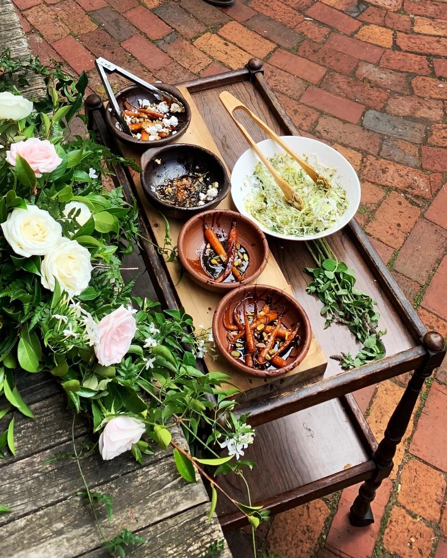 Event catering, empty wooden table with salad, multiple bowls with sauce and carrots, some greenery, and flowers on a brick patio.