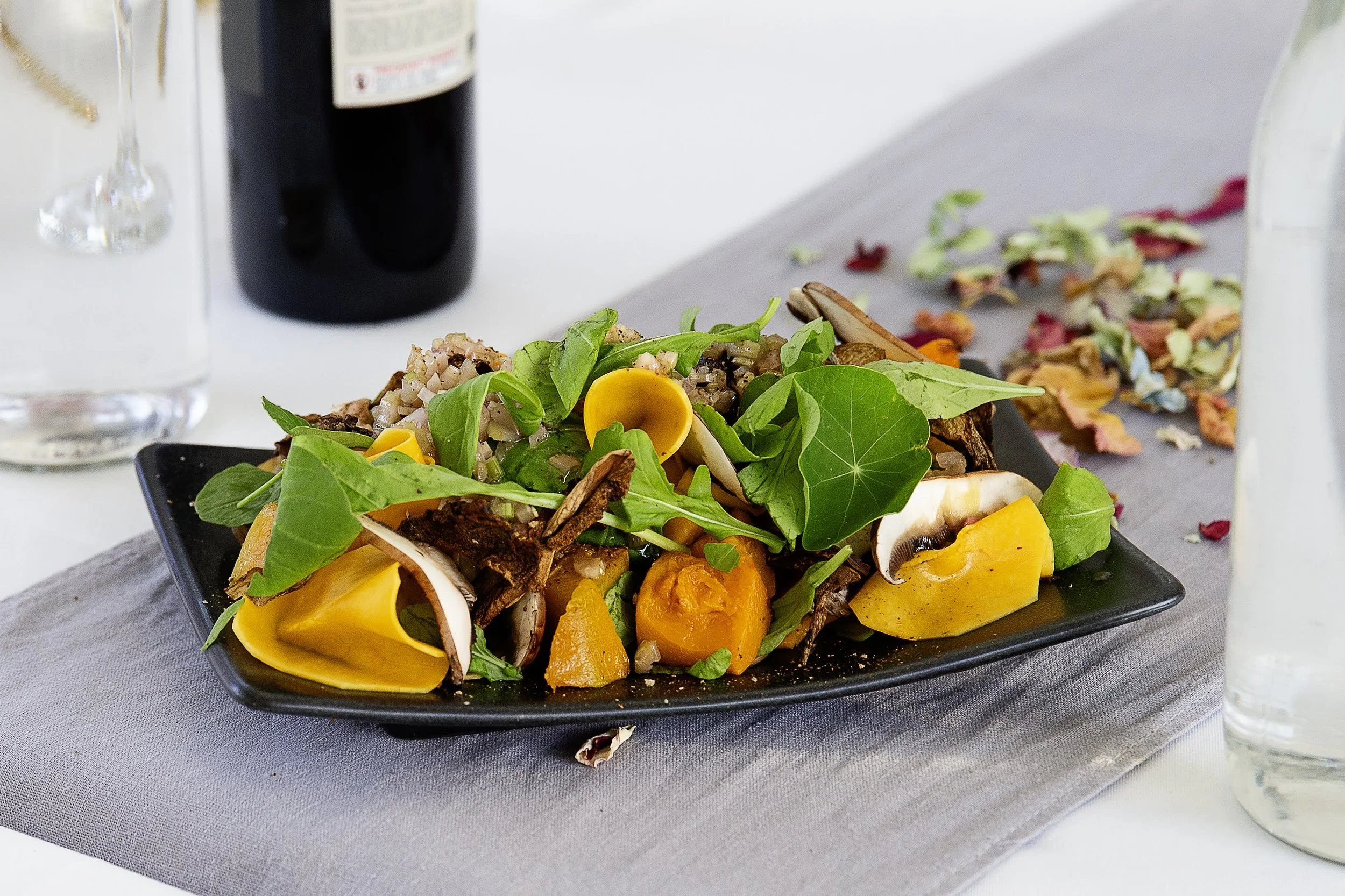 Fresh vegetable salad on a black plate with mixed greens, mushrooms, yellow peppers, tomatoes, and herbs, placed on a gray placemat with a glass of water and a bottle in the background.