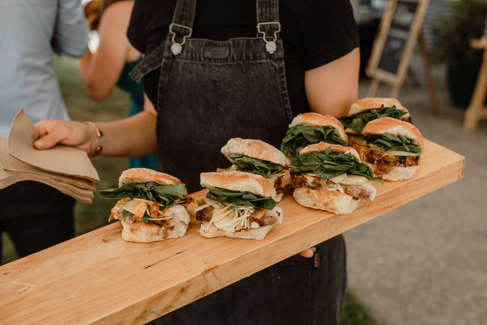 Person in black shirt and apron holding a wooden tray with several sandwiches at an outdoor event that has been catered.