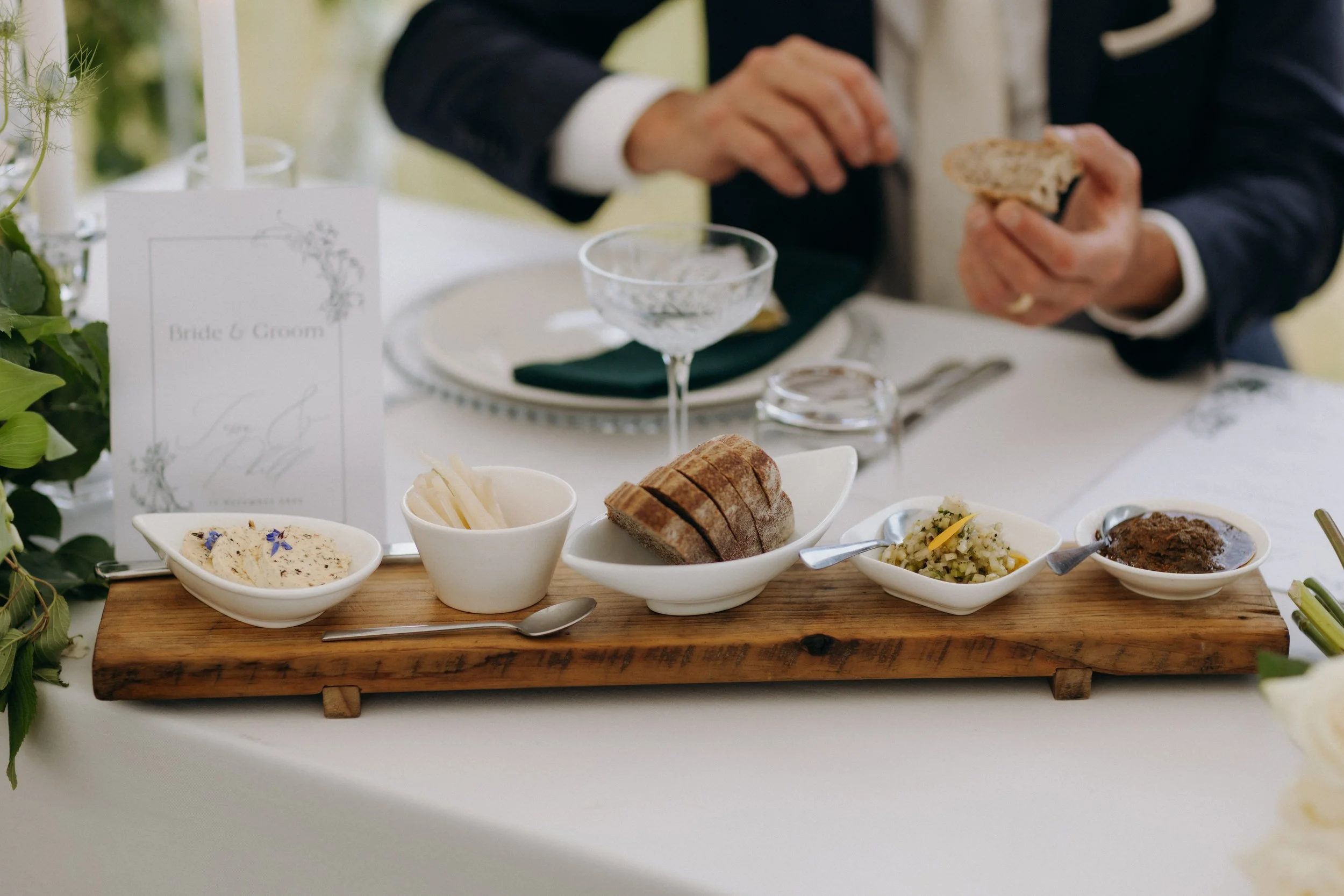A wooden serving board with various breads and spreads at a wedding reception table, with a wedding menu and a person in a tuxedo in the background.