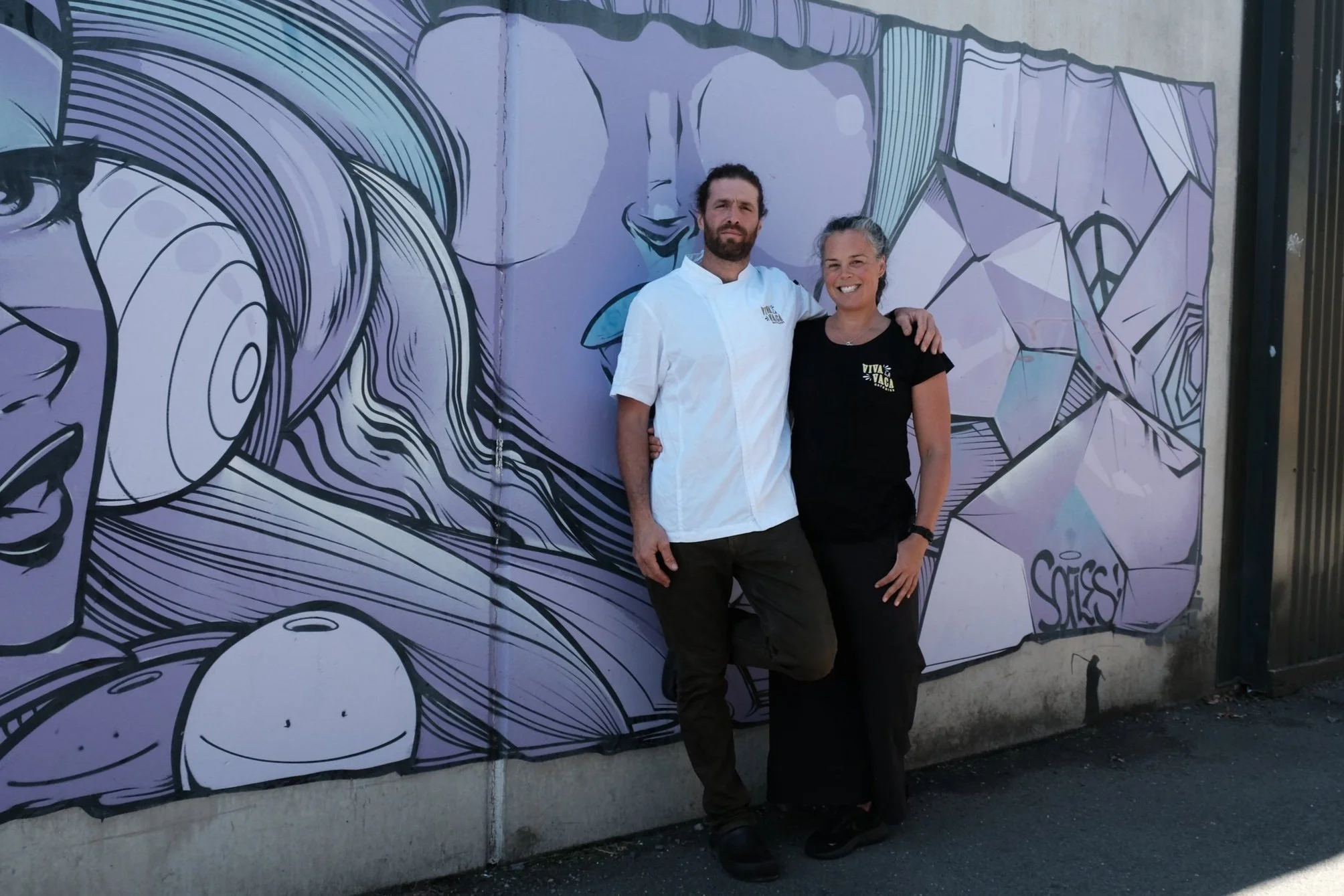 Caterers standing together, one man with a beard wearing a white chef's coat and a woman with gray hair wearing a black t-shirt. The mural features stylized faces and abstract patterns in shades of purple, blue, and black.