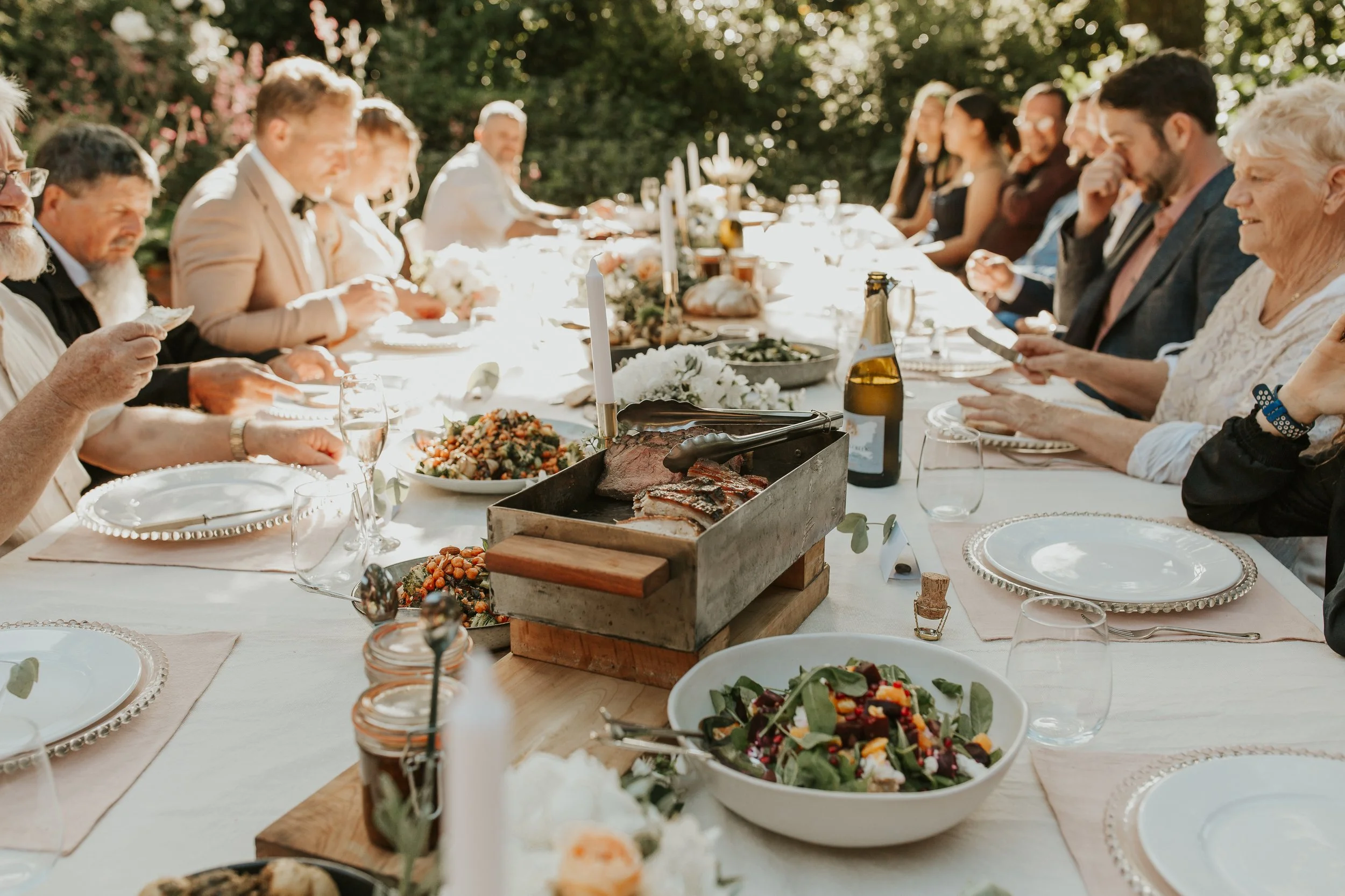 Family and friends gathered around a long outdoor dining table for a celebration, enjoying food and drinks in a lush garden setting.