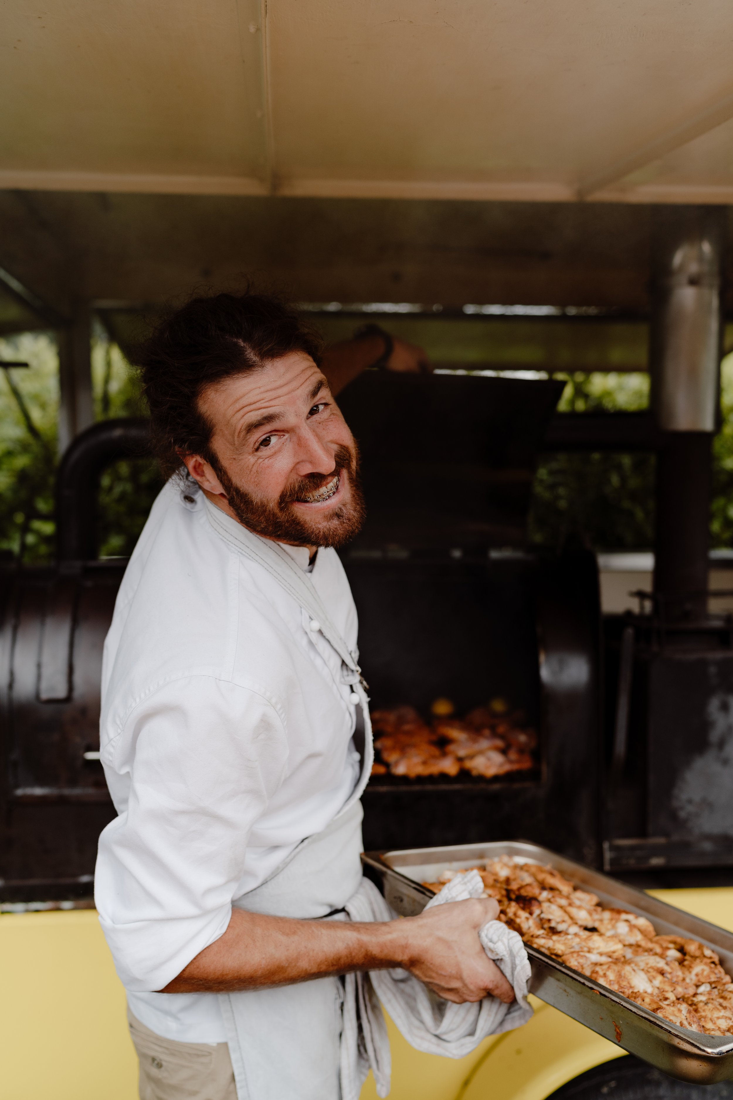 Man cooks meat on outdoor barbecue grill, holding tray of cooked meat, smiling at camera. He is the owner of the business, event caterer Matu.
