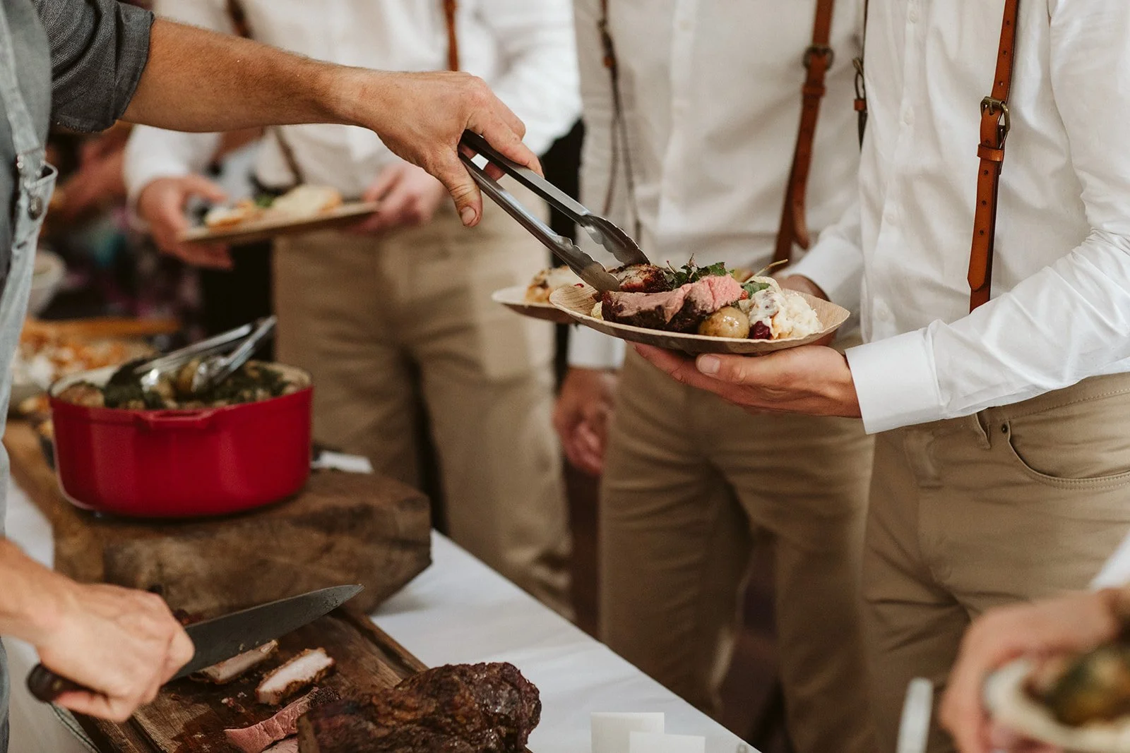 Person using tongs to serve food onto a plate at a buffet, with other people in line and food on the table. This is a catered event.