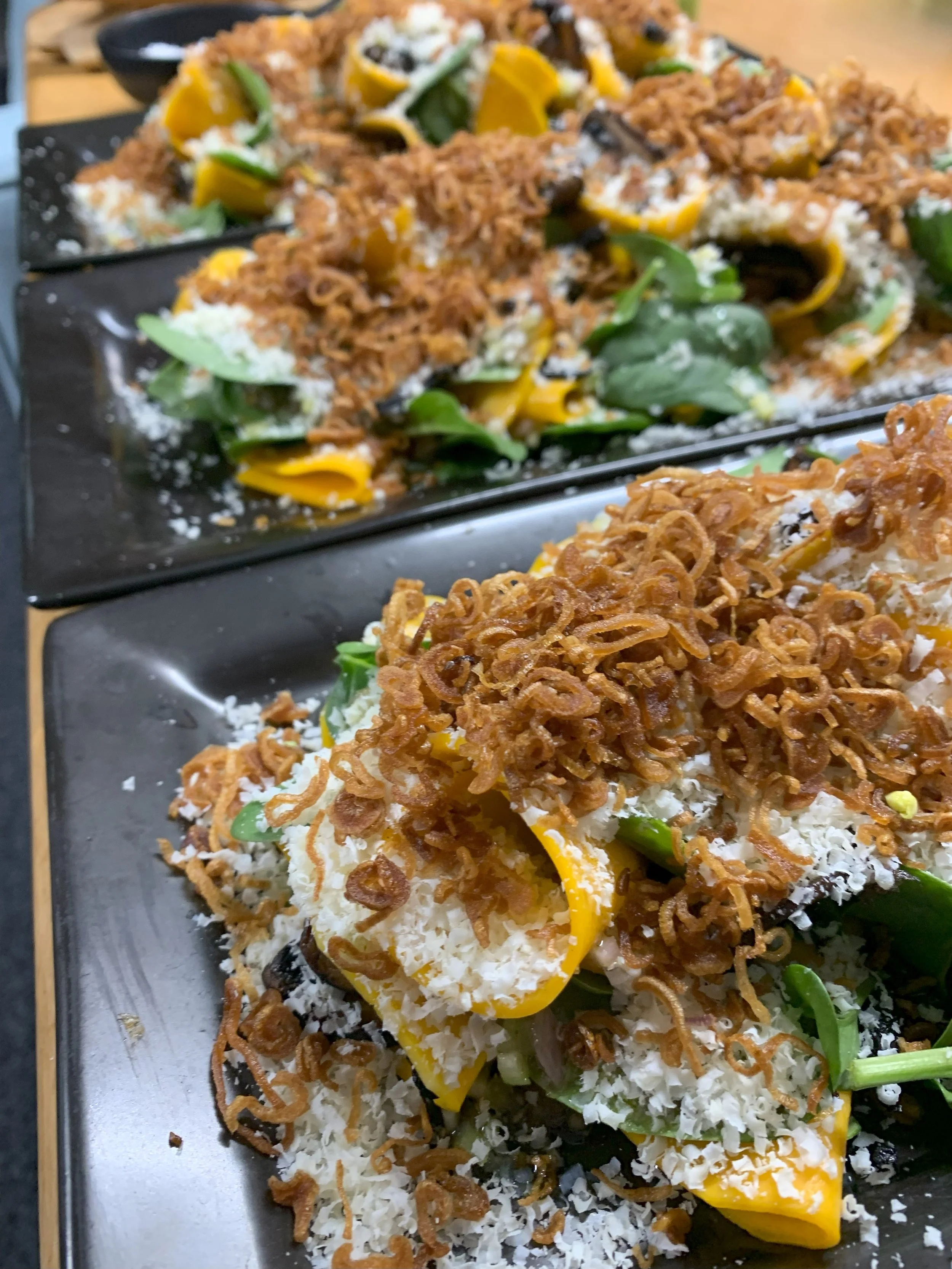 Two black plates of Israeli salad topped with fried onions, shredded cheese, yellow bell peppers, and green herbs on a wooden table.